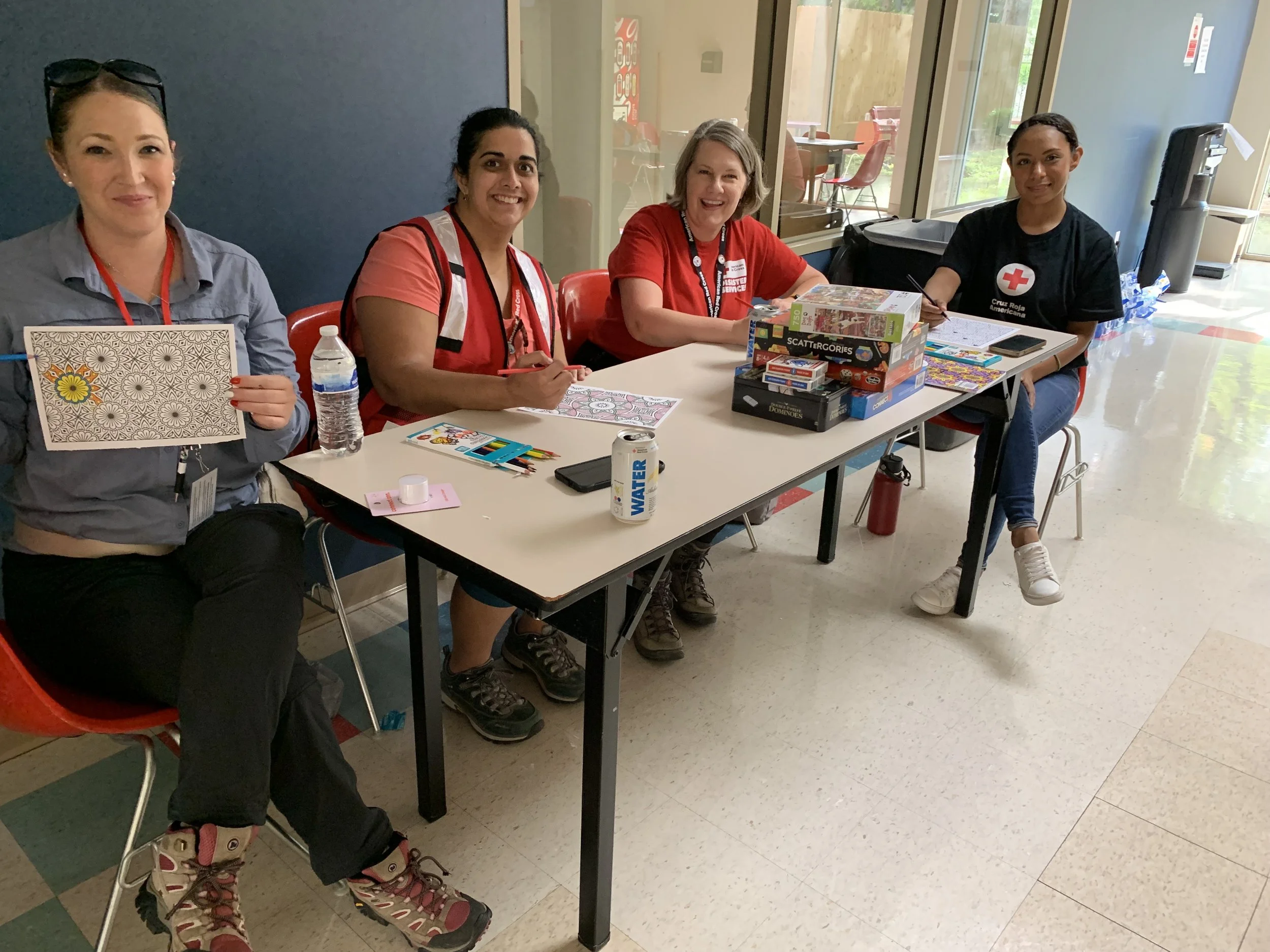 Group of four women sitting at a table, engaged in coloring and crafts, with board games on the table, in a community center setting.