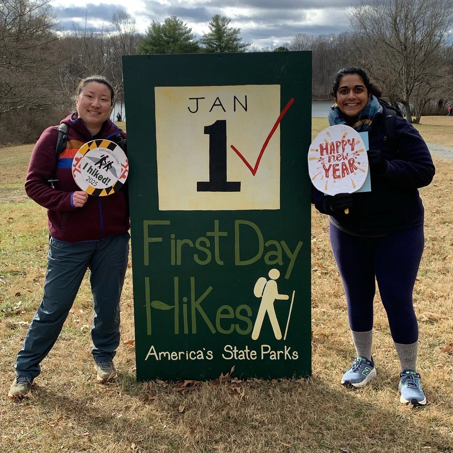 Two people standing beside a "First Day Hikes, America's State Parks" sign on January 1, holding celebration signs. It is an outdoor scene with trees in the background.