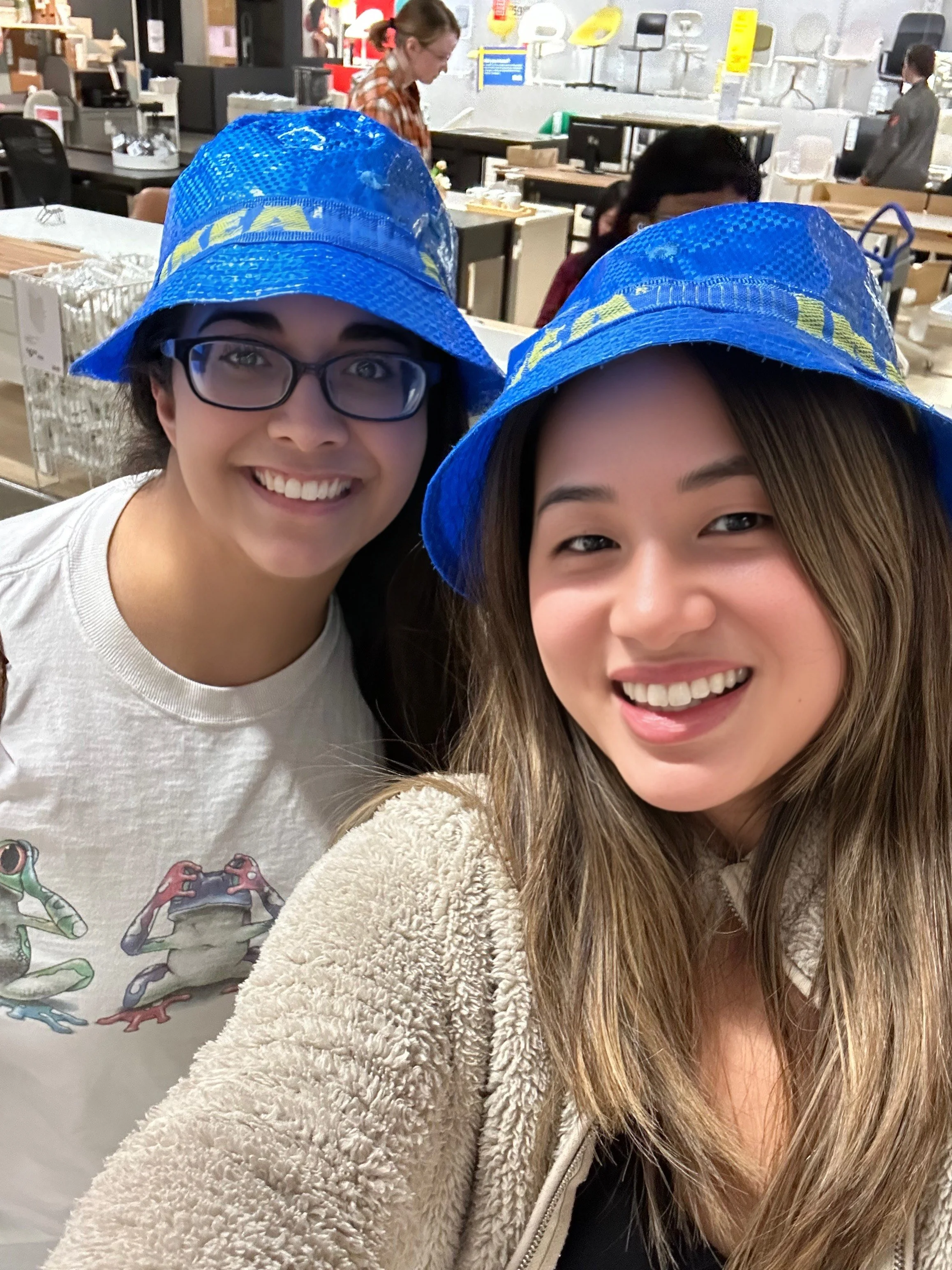 Two people wearing blue IKEA hats, smiling in a store setting with shelves and chairs in the background.