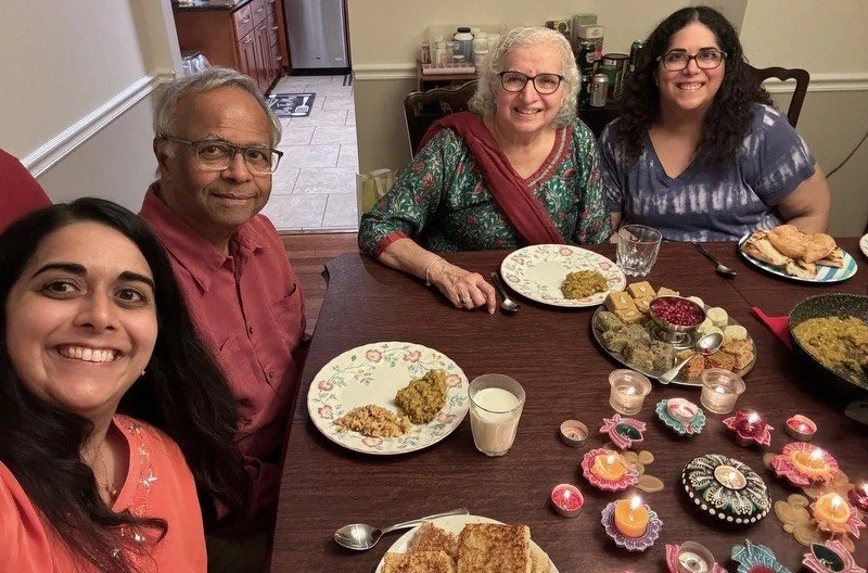 Candidate's family celebrating Diwali sitting around a dining table with food, candles, and decorative items, smiling for a photo. The table has plates with food, bread, and drinks. The room is warmly lit and looks like a home setting.