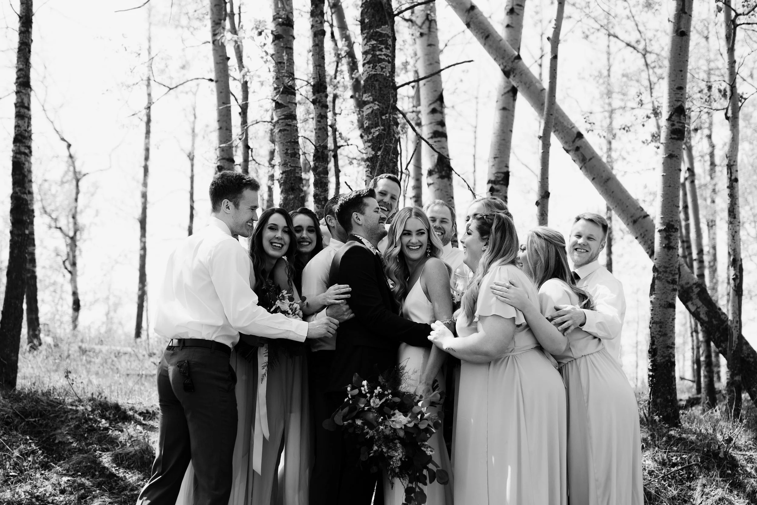 A joyful wedding party surrounds the bride and groom in a playful group hug among tall, sunlit birch trees. The black-and-white photo captures genuine laughter, candid emotion, and the natural beauty of an outdoor forest setting. This fun, heartfelt 