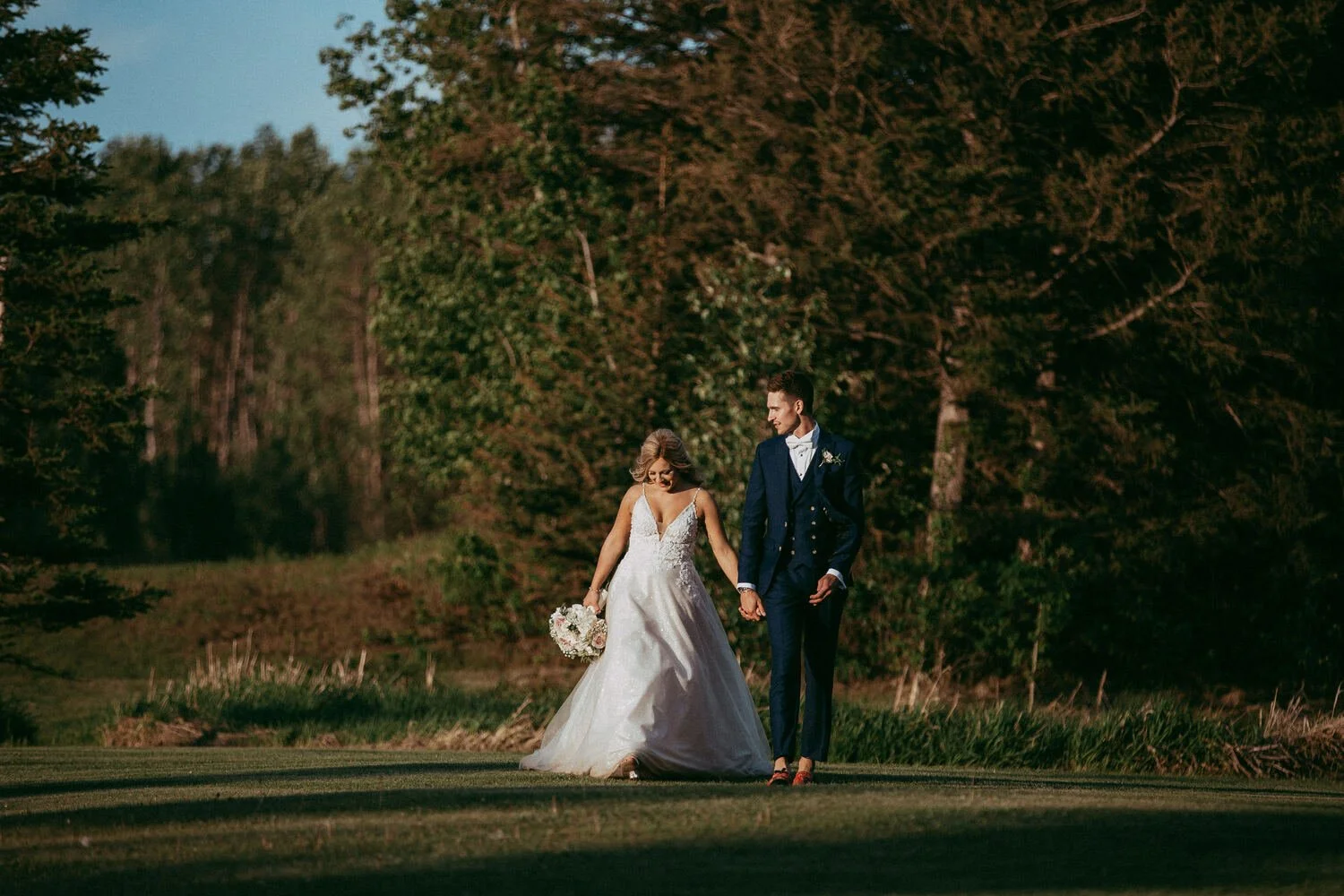A bride and groom walk hand-in-hand through the golden evening light at Pine and Pond Wedding Venue, surrounded by lush trees and peaceful countryside views. 