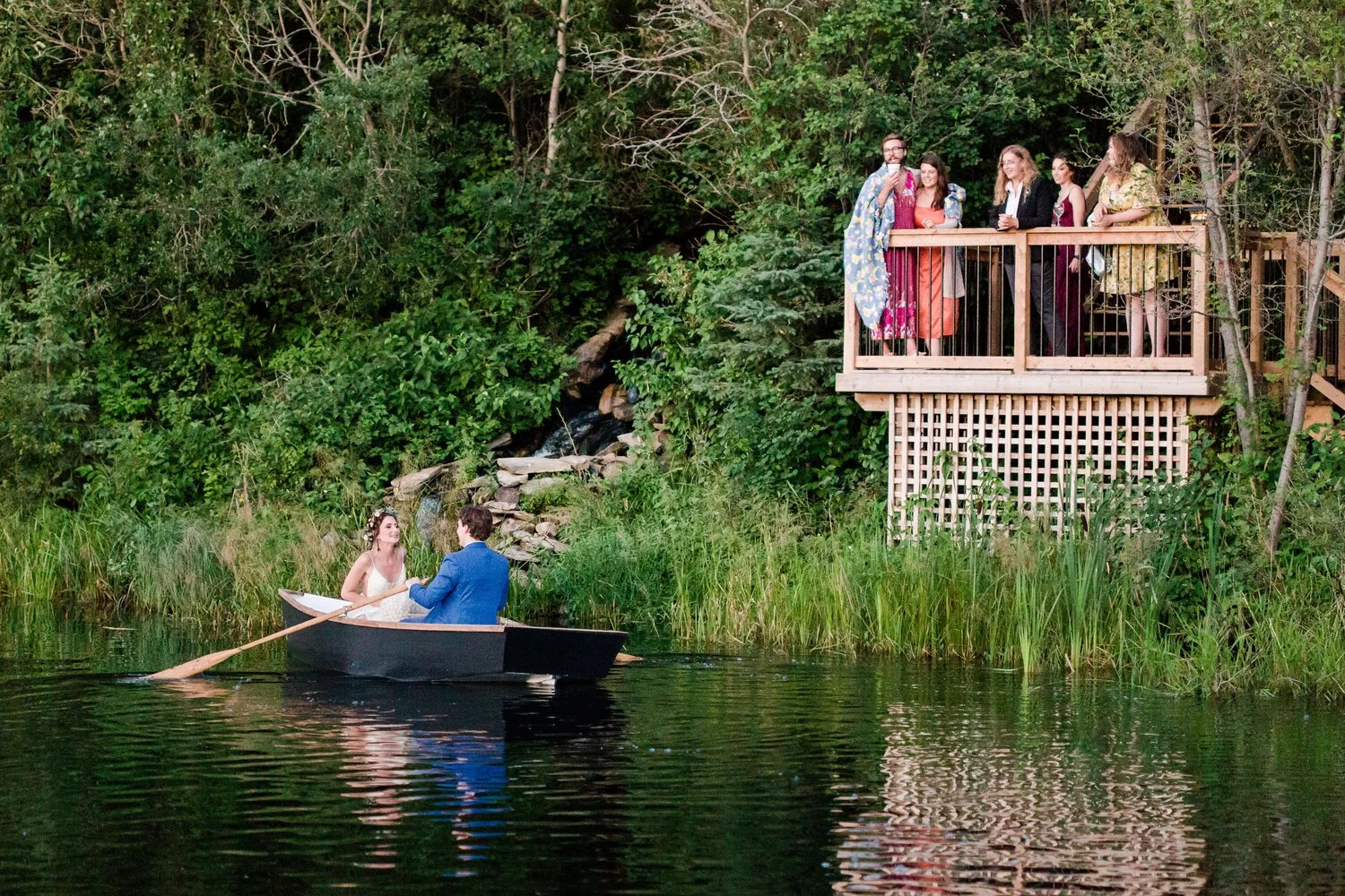 Bride and groom sitting in a rowboat on the pond as guests watch from the viewing deck at Pine and Pond