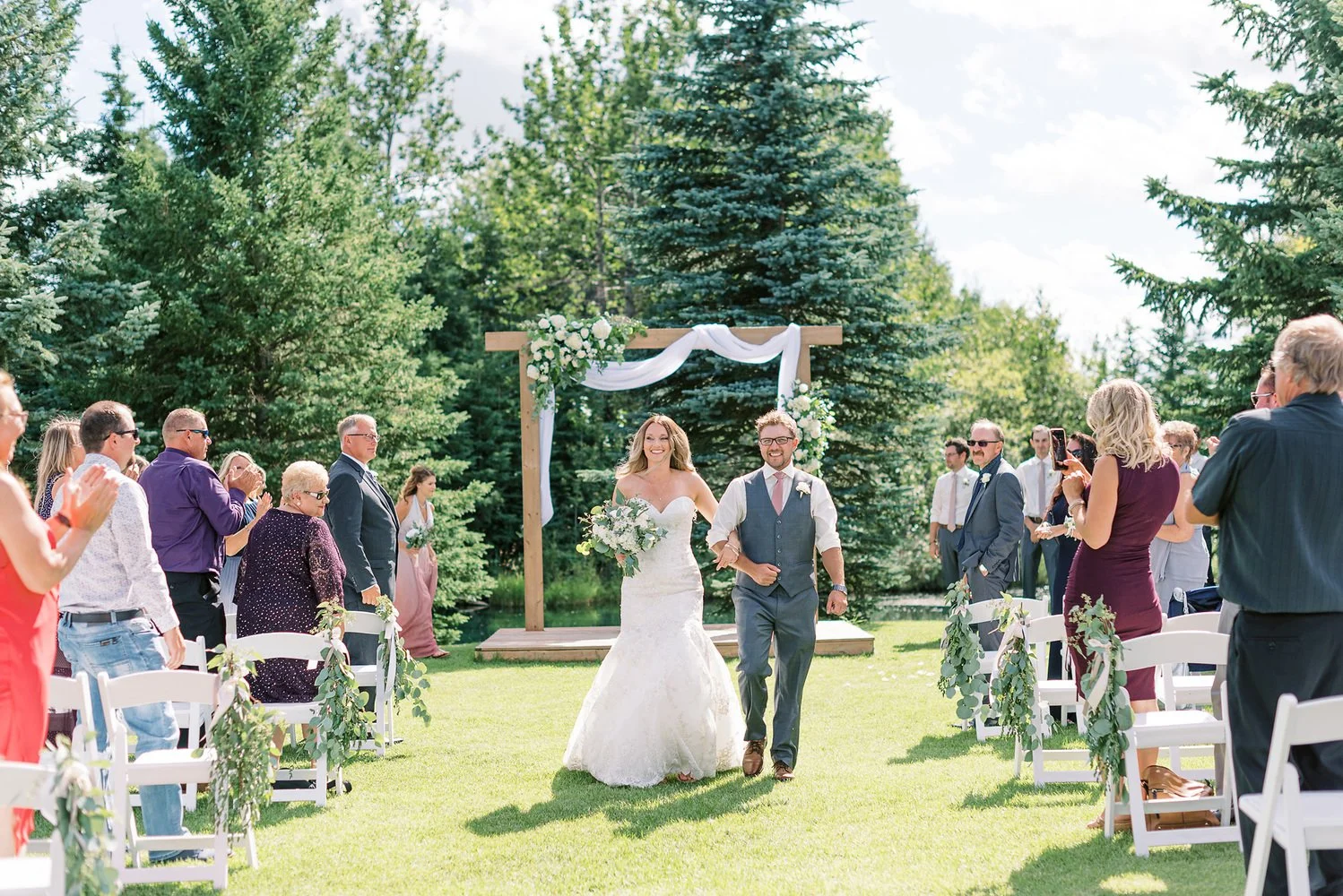 Outdoor wedding ceremony at Pine and Pond in Alberta with guests holding umbrellas during light rain, surrounded by trees