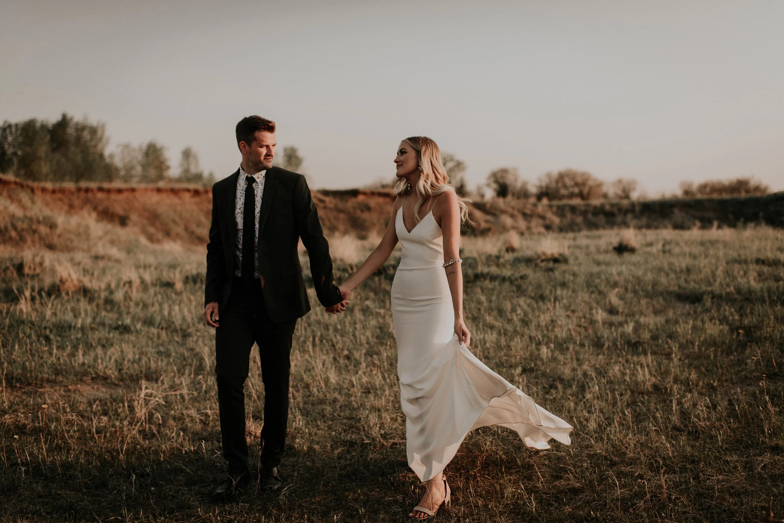 A bride and groom walk hand in hand through a golden field at sunset, creating a romantic and cinematic wedding portrait. The bride’s sleek white gown flows behind her in the warm evening breeze, while the groom looks toward her with a soft, natural 