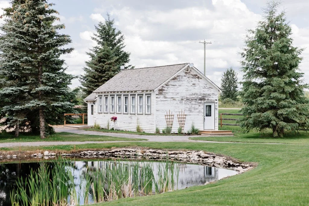 Exterior of the historic white bridal cottage at Pine and Pond Wedding Venue, surrounded by mature trees and overlooking the pond — a charming backdrop for wedding detail photos.