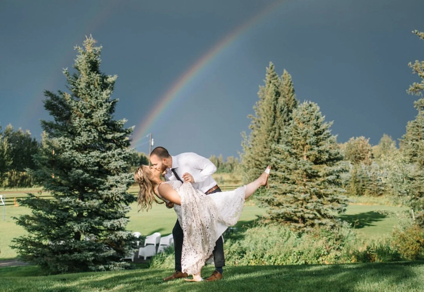 A bride and groom share a romantic dip kiss on a lush green lawn with a vibrant rainbow arching across the stormy sky behind them. Surrounded by tall evergreen trees and dramatic lighting, the scene feels magical and spontaneous. The bride’s lace dre