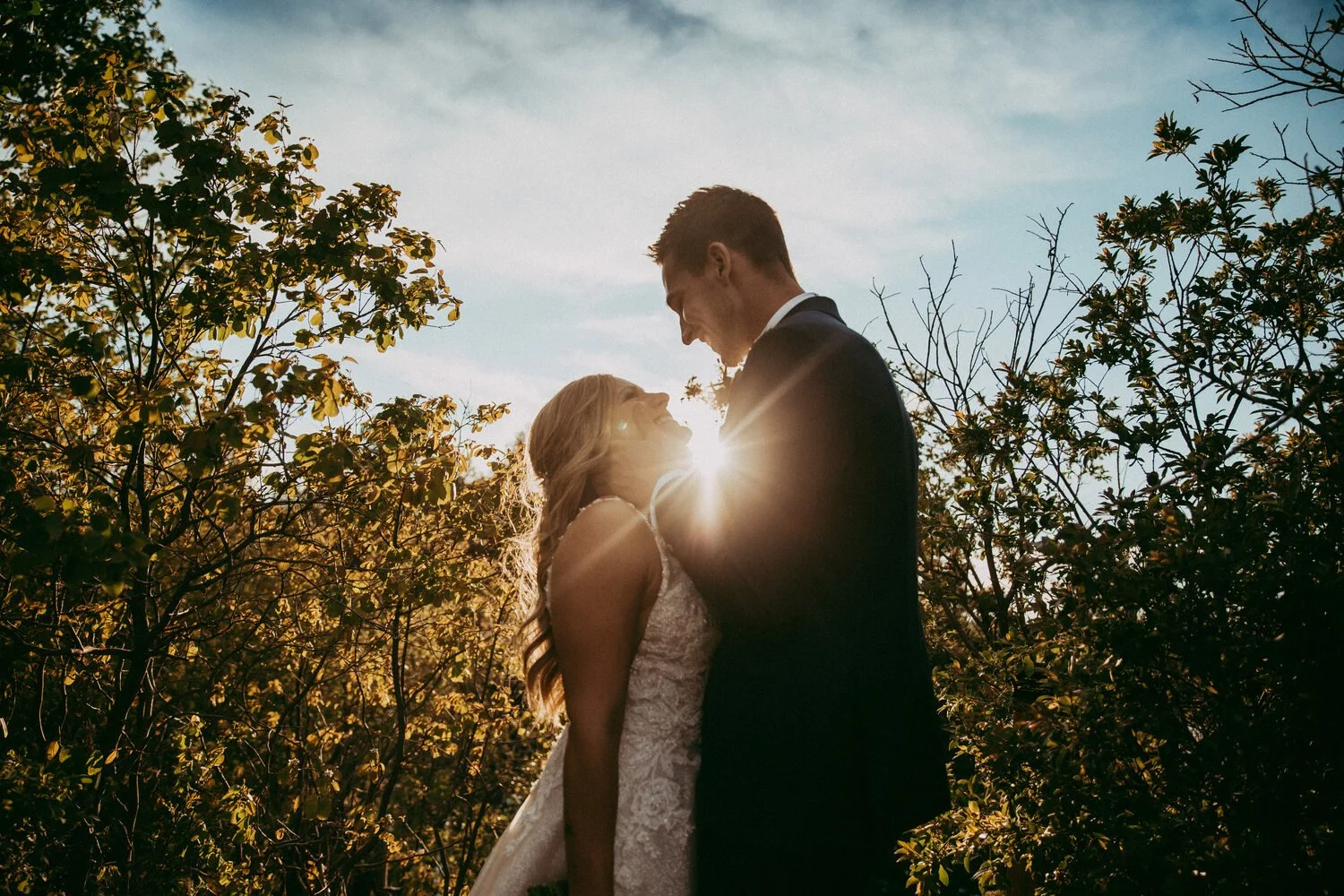 A bride and groom share an intimate embrace at sunset, surrounded by glowing foliage as warm golden light shines through the trees. The sunburst peeks between them, illuminating the bride’s lace gown and the groom’s suit while they smile at each othe