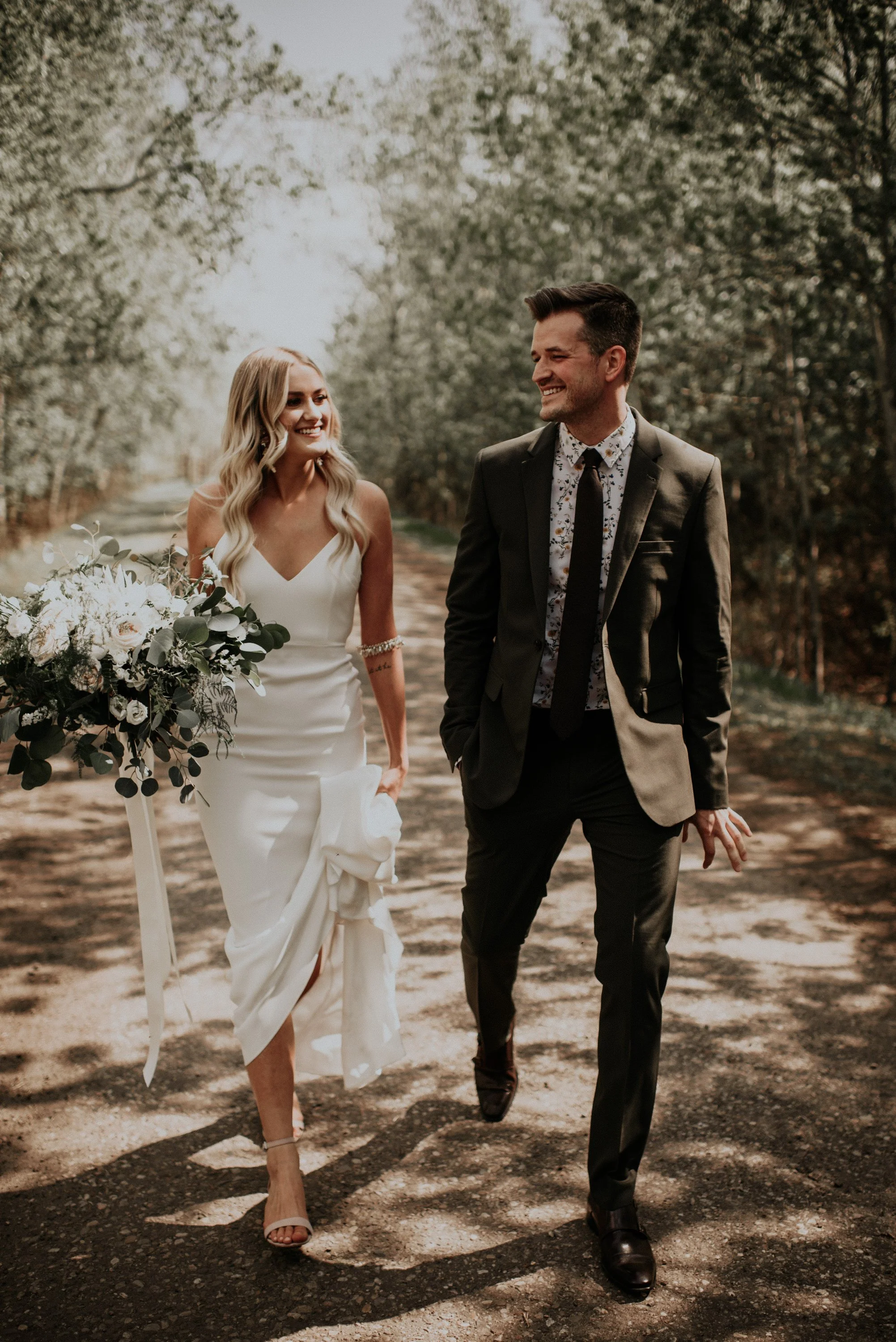 A bride and groom walk hand in hand down a tree-lined path, smiling warmly at each other during their outdoor wedding. The bride carries a lush bouquet of white flowers and greenery, while her sleek, modern gown contrasts beautifully with the natural