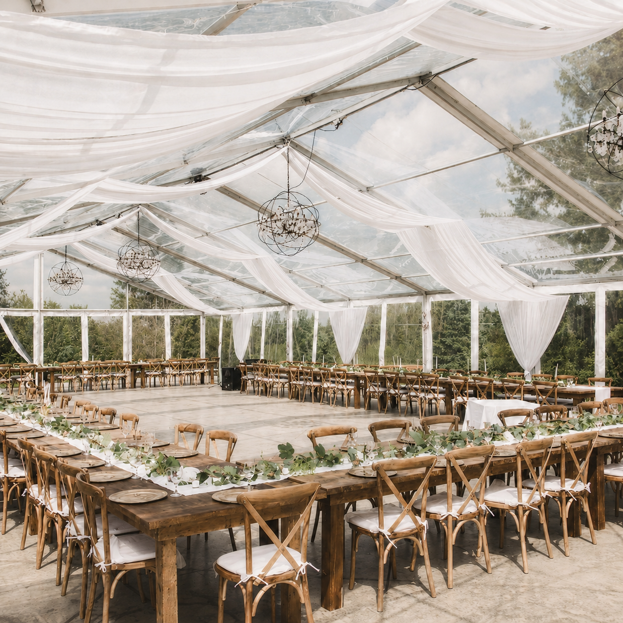 Outdoor wedding reception in a white tent with wooden tables and chairs, decorated with greenery and chandeliers.