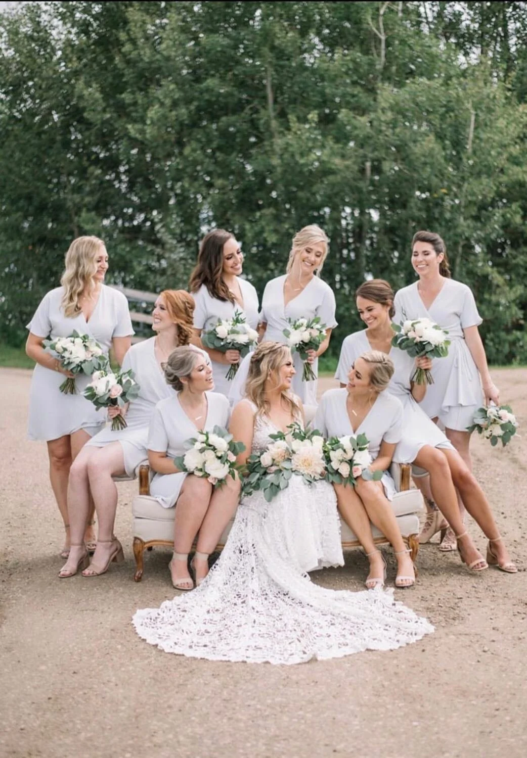 A bride sits with her bridesmaids on a vintage loveseat along a rustic country road, surrounded by lush green trees. The bridal party wears matching light blue dresses and holds classic white and greenery bouquets, creating a fresh, elegant, and cohe