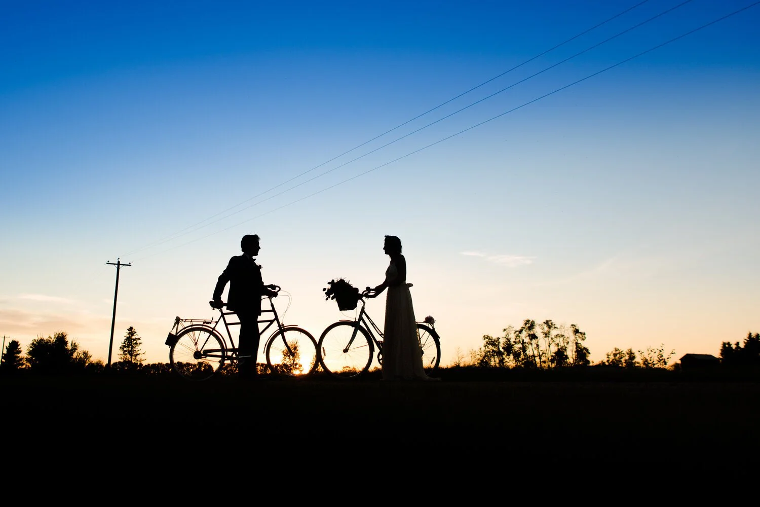 Silhouetted bride and groom standing with bicycles at sunset at Pine and Pond Wedding Venue, sharing a quiet romantic moment against the evening sky in Central Alberta.