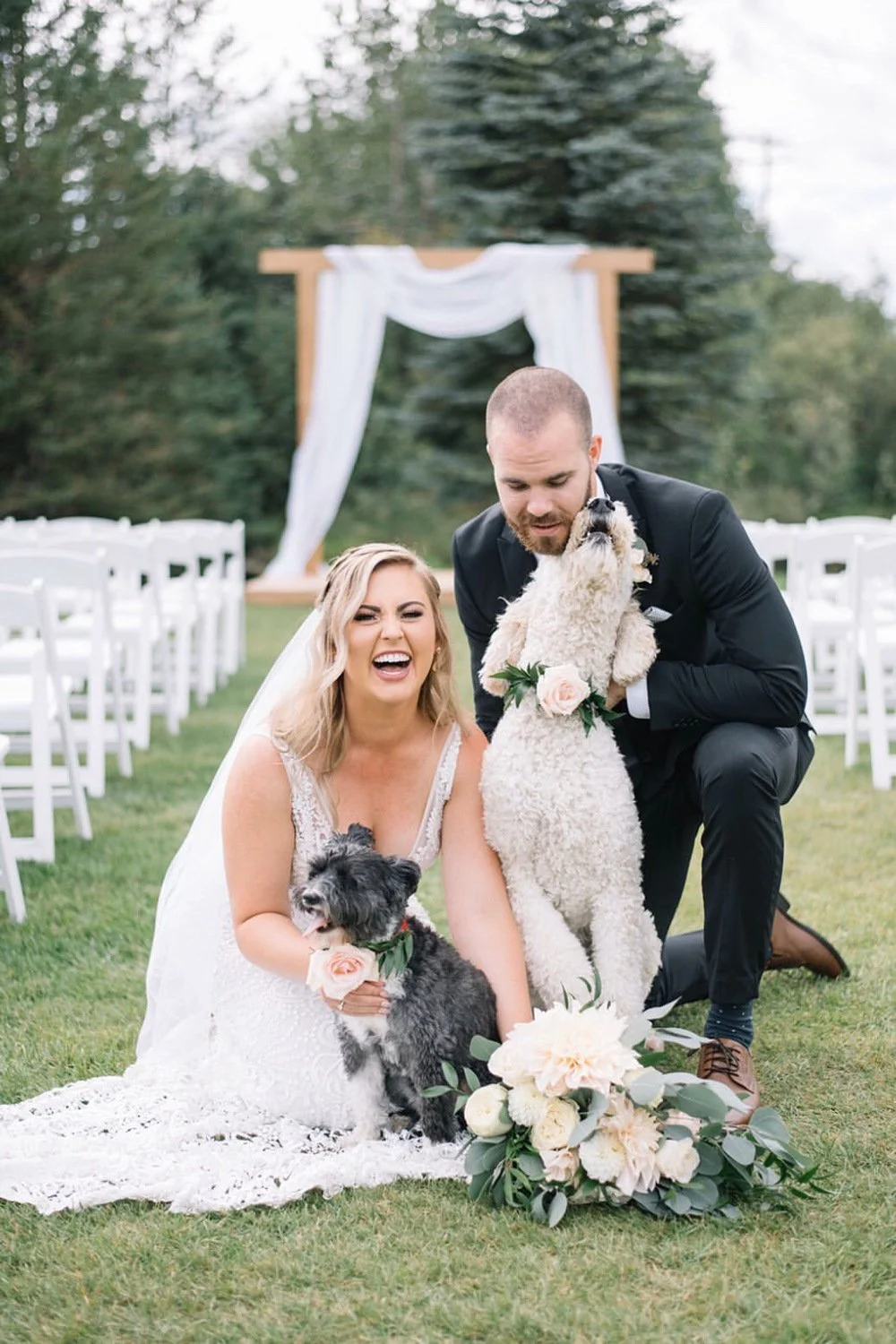 A bride and groom share a joyful moment with their two dogs during an outdoor wedding ceremony. The couple kneels on the grass in front of a wooden arbor draped with white fabric, laughing as their pups—wearing floral collars—snuggle close. The bride