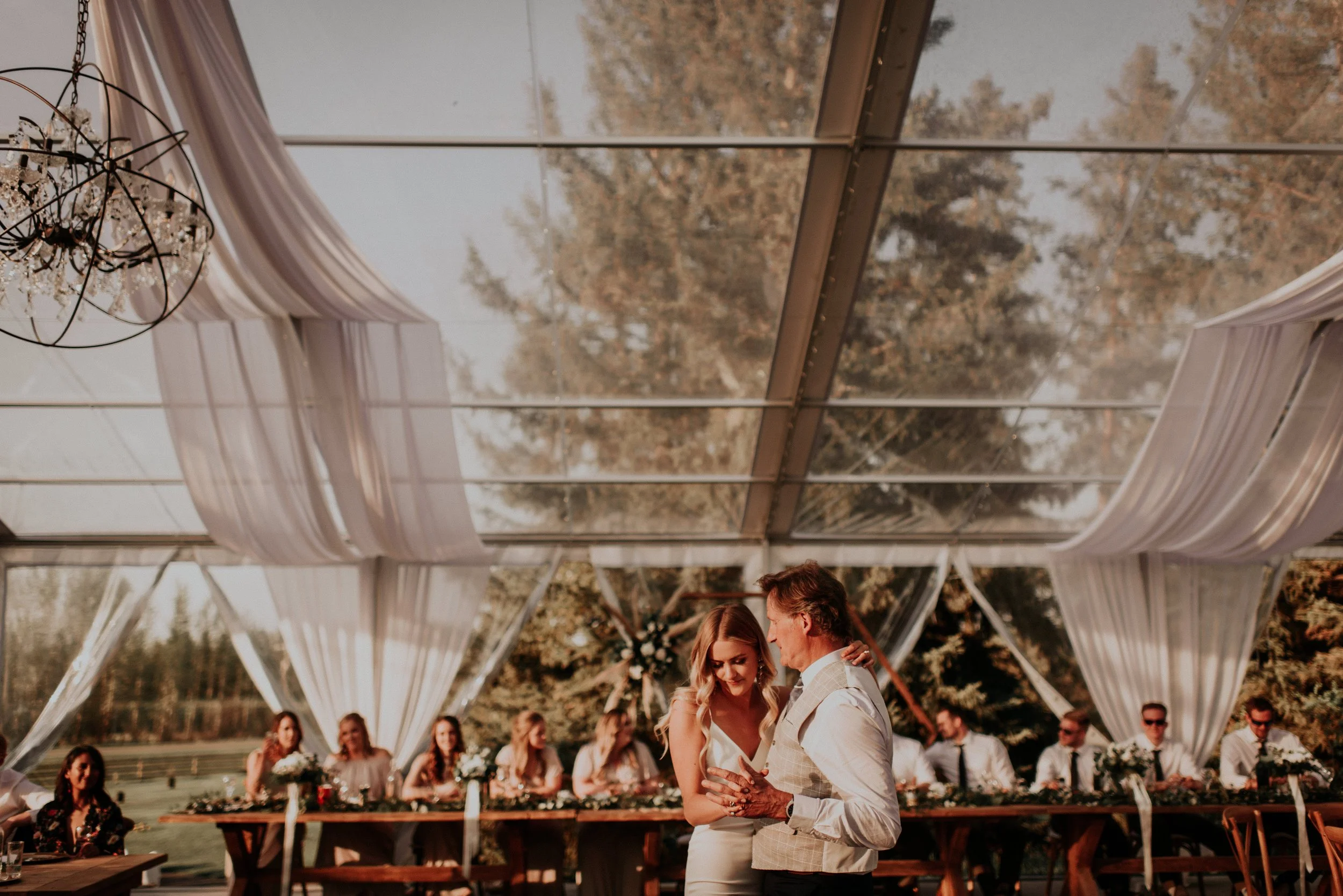 A bride shares an emotional father–daughter dance beneath a stunning clear-top wedding tent draped with soft white fabric. Warm golden-hour light filters through the transparent roof, highlighting the bride’s elegant gown and her father’s heartfelt e