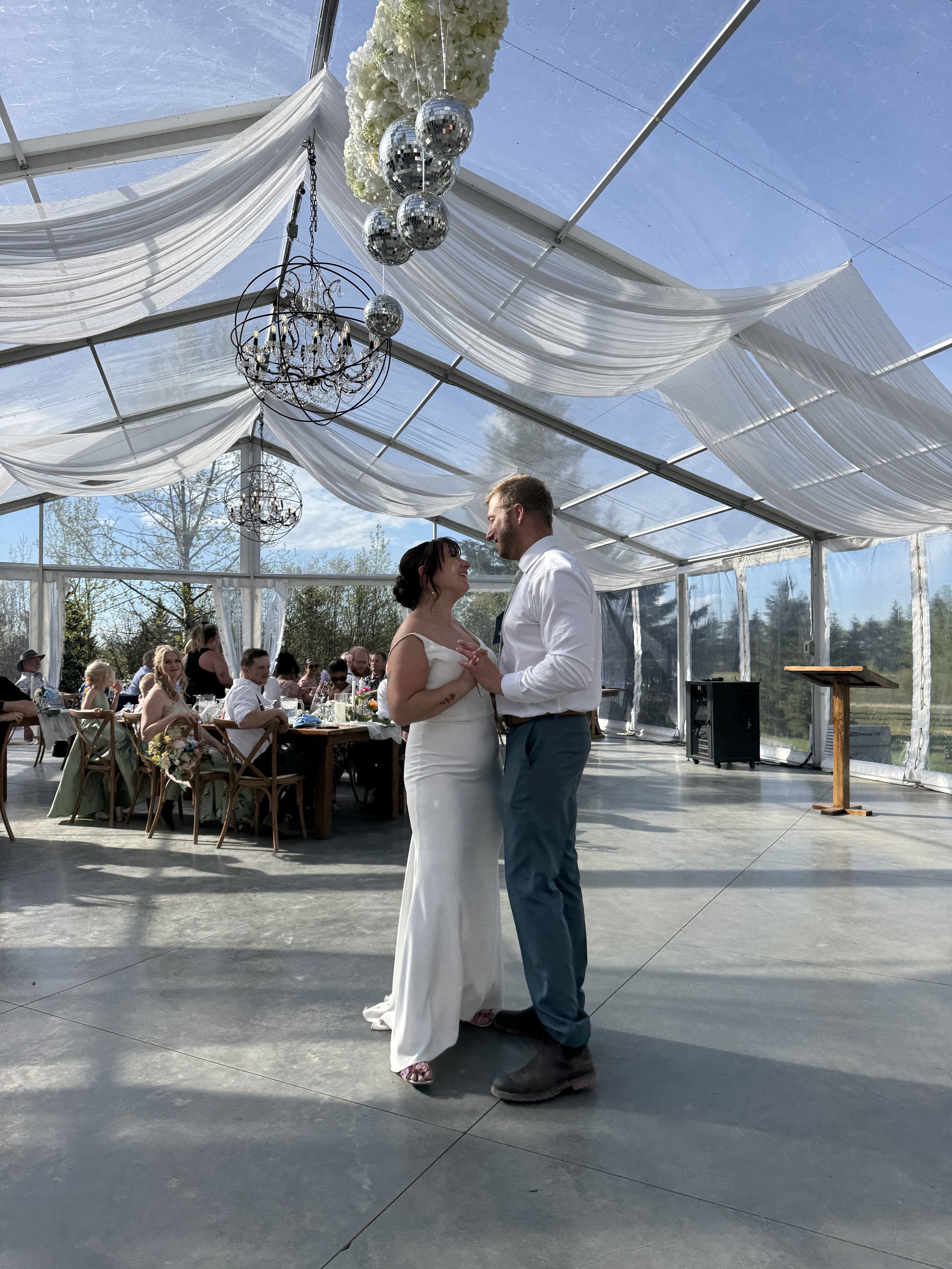A newly married couple shares their first dance beneath chandeliers and flowing drapery inside Pine and Pond's elegant clear tent wedding venue in Central Alberta.