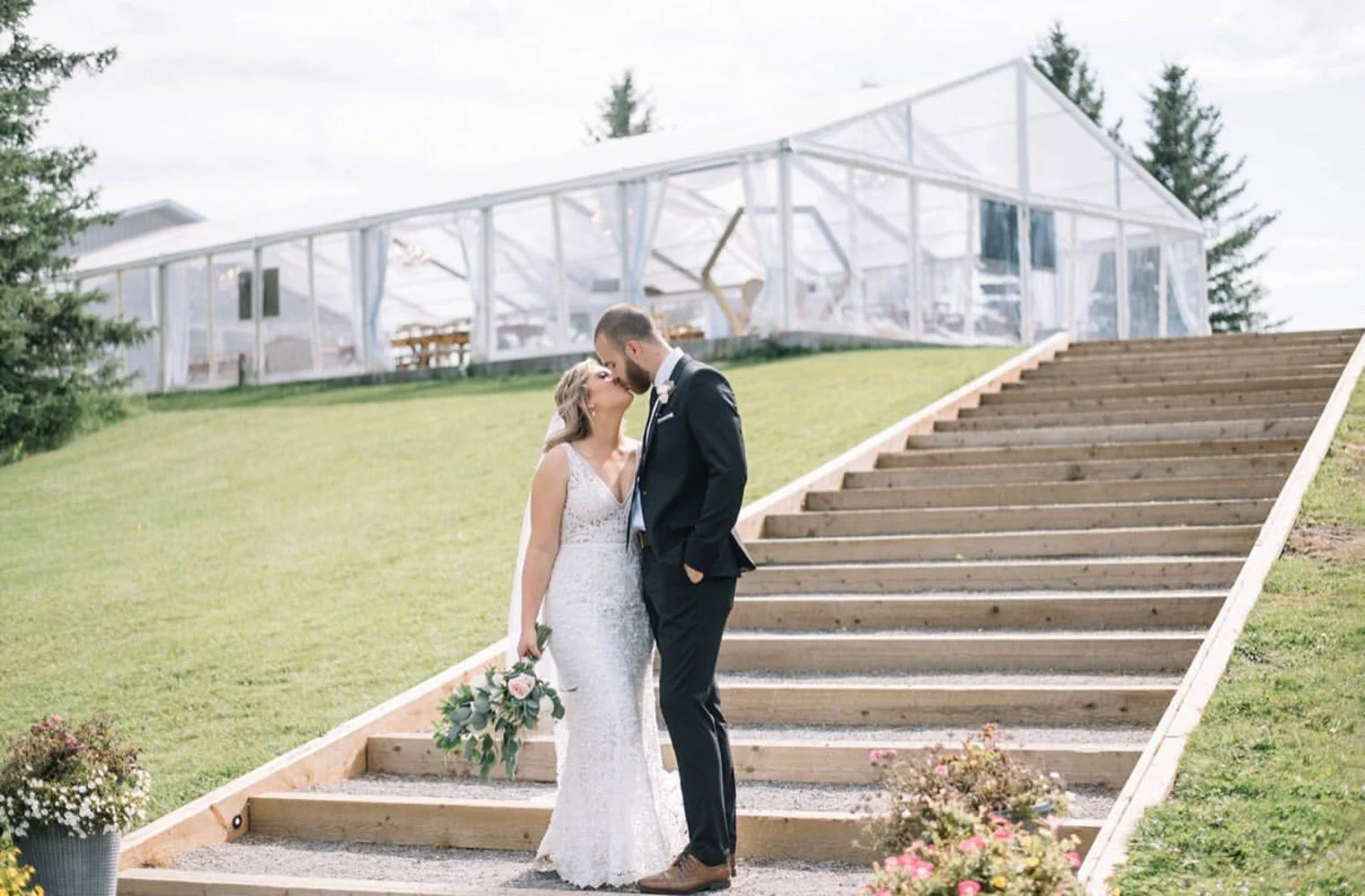 A bride and groom share a sweet kiss at the base of a wide wooden staircase leading up to a stunning clear-top wedding tent. The bride’s lace gown and pastel bouquet complement the bright, airy backdrop, while the groom’s classic dark suit adds elega