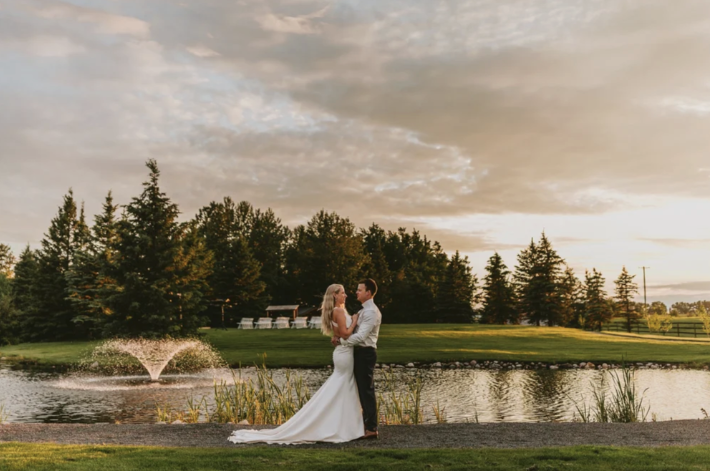 Bride and groom embracing beside the pond at sunset at Pine and Pond Wedding Venue, with a water fountain and golden evening light reflecting across the grounds