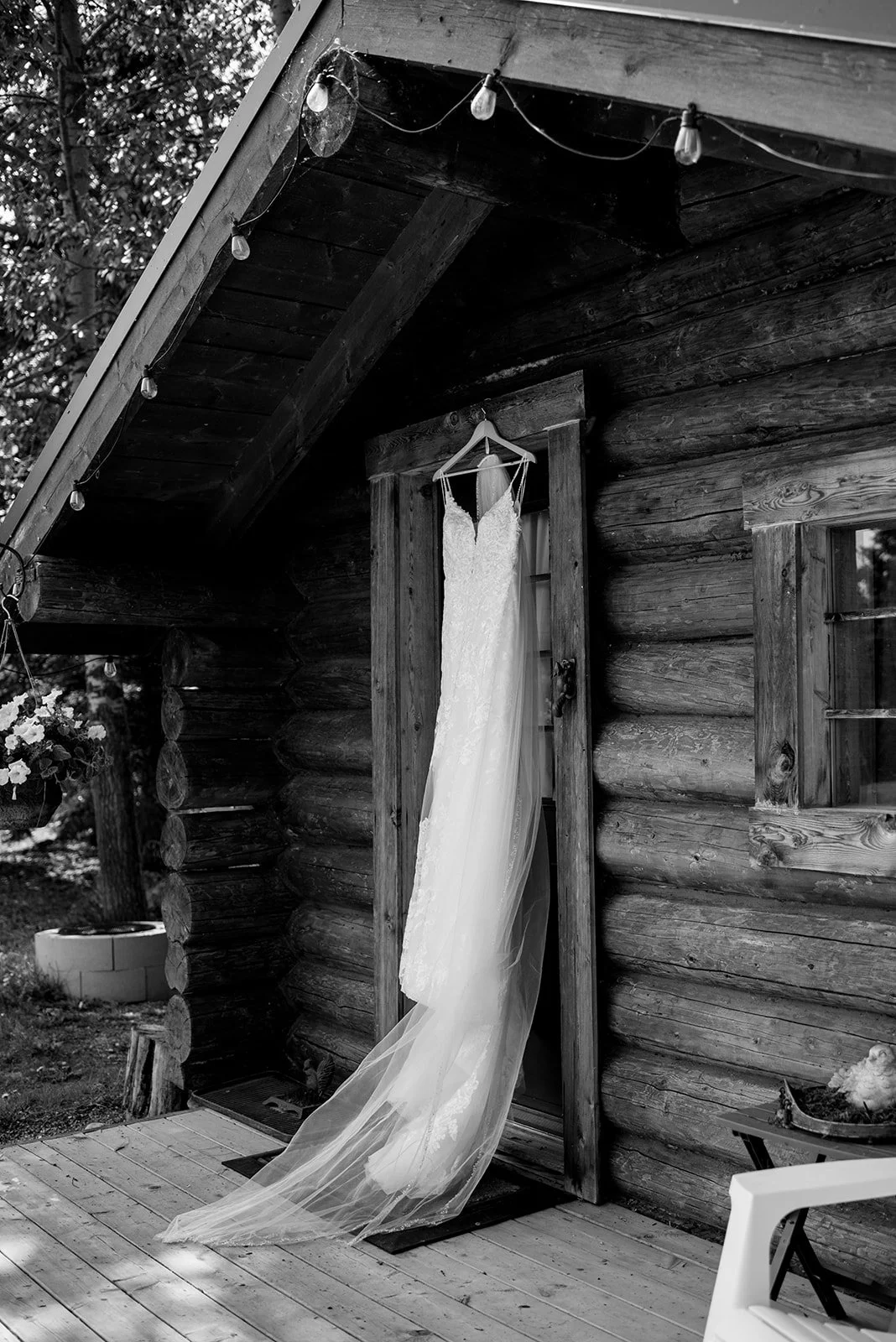 A lace wedding dress hangs elegantly on the door of a rustic log cabin, its long tulle train cascading onto the wooden porch. Soft natural light highlights the cabin’s weathered wood texture, creating a charming, nature-inspired backdrop. This black-