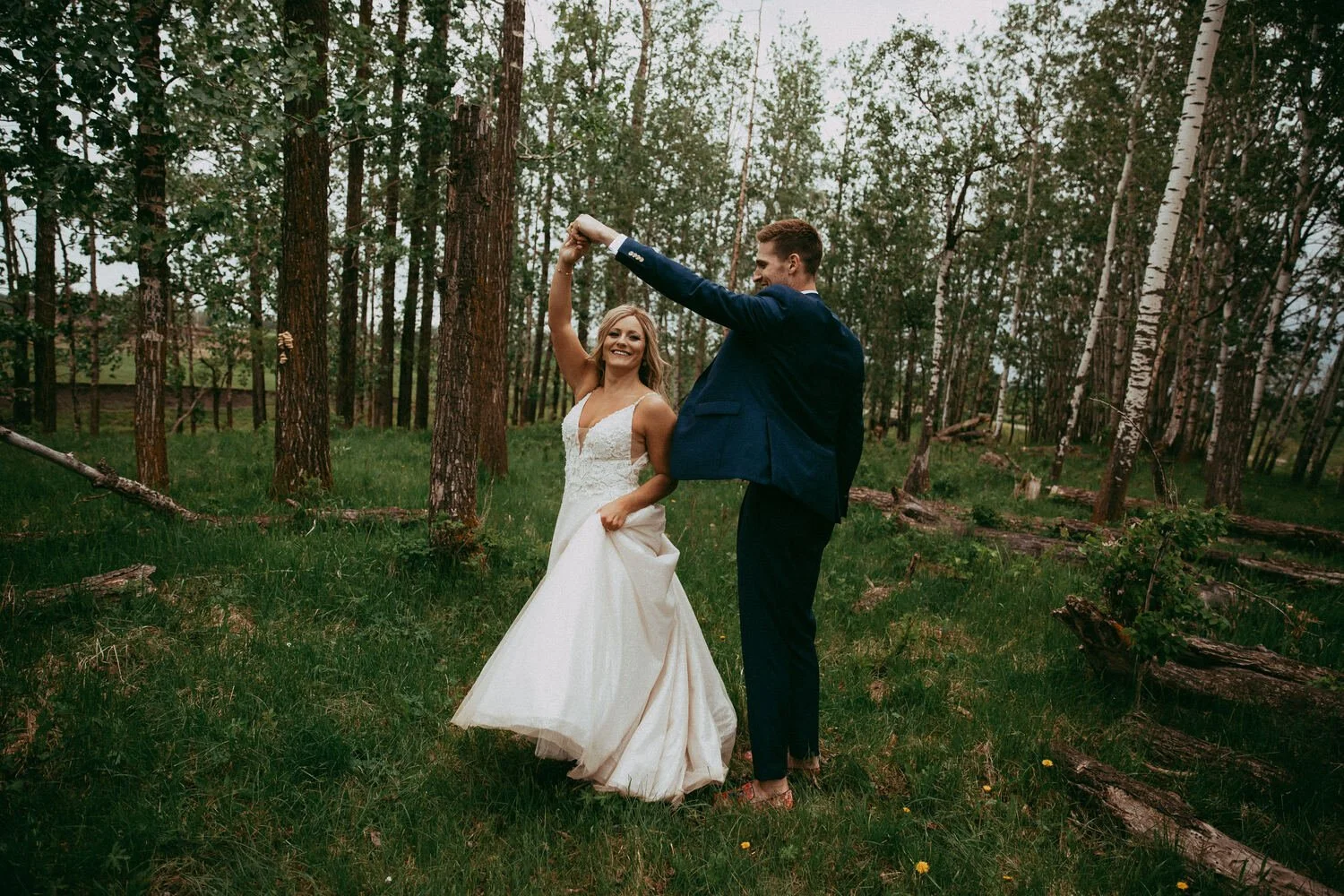 A bride and groom twirl together in a lush forest clearing, surrounded by tall trees, fallen logs, and vibrant green grass. The bride’s flowing gown moves beautifully as she spins, while the groom smiles and leads her in a playful dance. This candid 