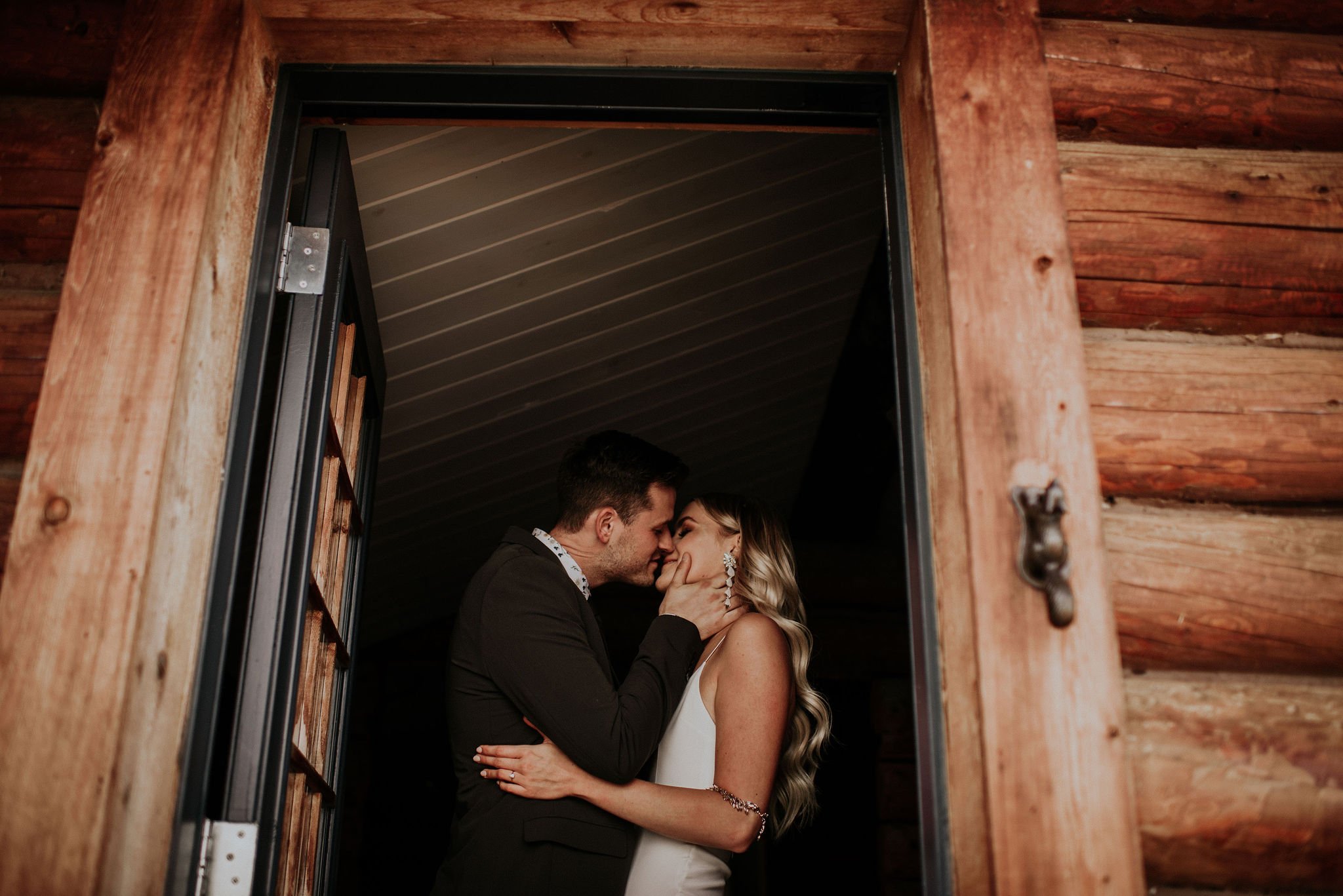 A bride and groom share a tender kiss in the doorway of a rustic log cabin, framed by warm wooden textures and soft indoor lighting. The groom gently holds the bride’s face as she smiles, capturing an intimate, emotional moment. Her long, wavy hair a