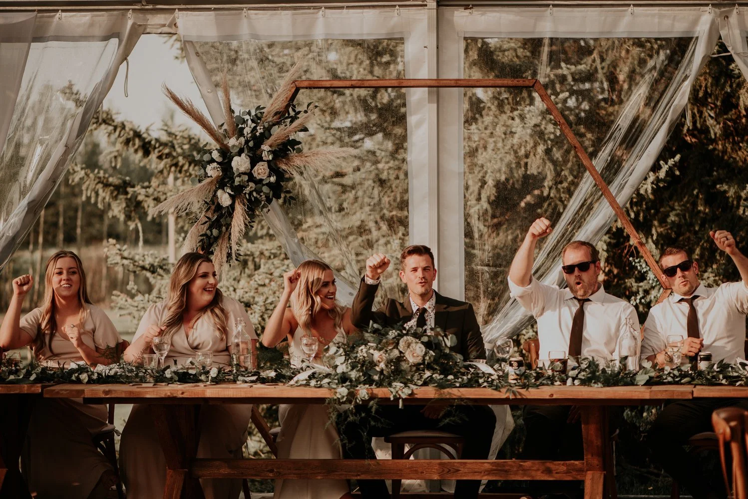 A lively wedding head table celebration takes place inside a clear-top reception tent, with the bride, groom, bridesmaids, and groomsmen raising their fists in joy. The long wooden harvest table is decorated with lush greenery, white roses, and elega