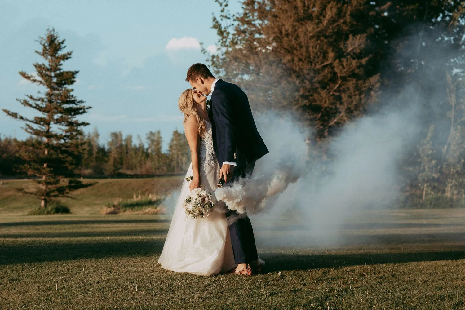 Amid the sweeping green landscape of Pine & Pond, this couple shares a tender kiss as soft clouds of celebratory smoke drift around them. Bathed in warm evening light, the moment feels beautifully cinematic—playful, romantic, and full of personality.