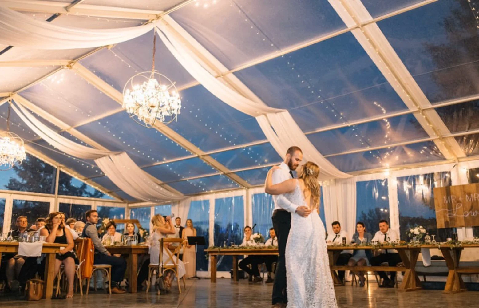 A bride and groom share their magical first dance under a clear-top wedding tent glowing with chandeliers, draped white fabric, and twinkling string lights. Guests look on from rustic wooden tables as the evening sky deepens to blue, creating a roman