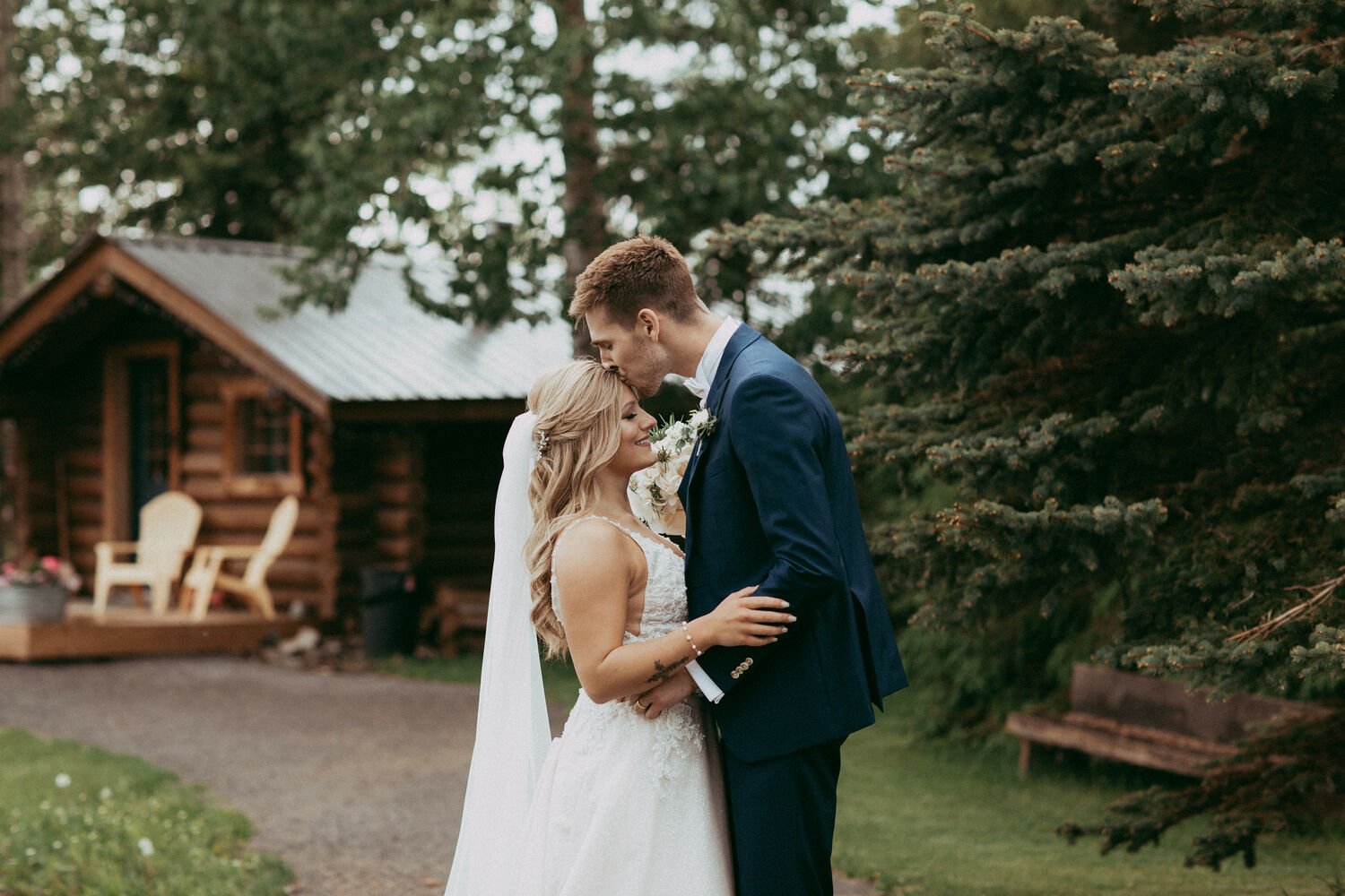 This tender moment captures the bride and groom sharing a soft embrace along the tree-lined path leading to the charming log cabin at Pine & Pond Wedding Venue in Central Alberta.  Perfect for couples who love rustic elegance, intimate portraits, and