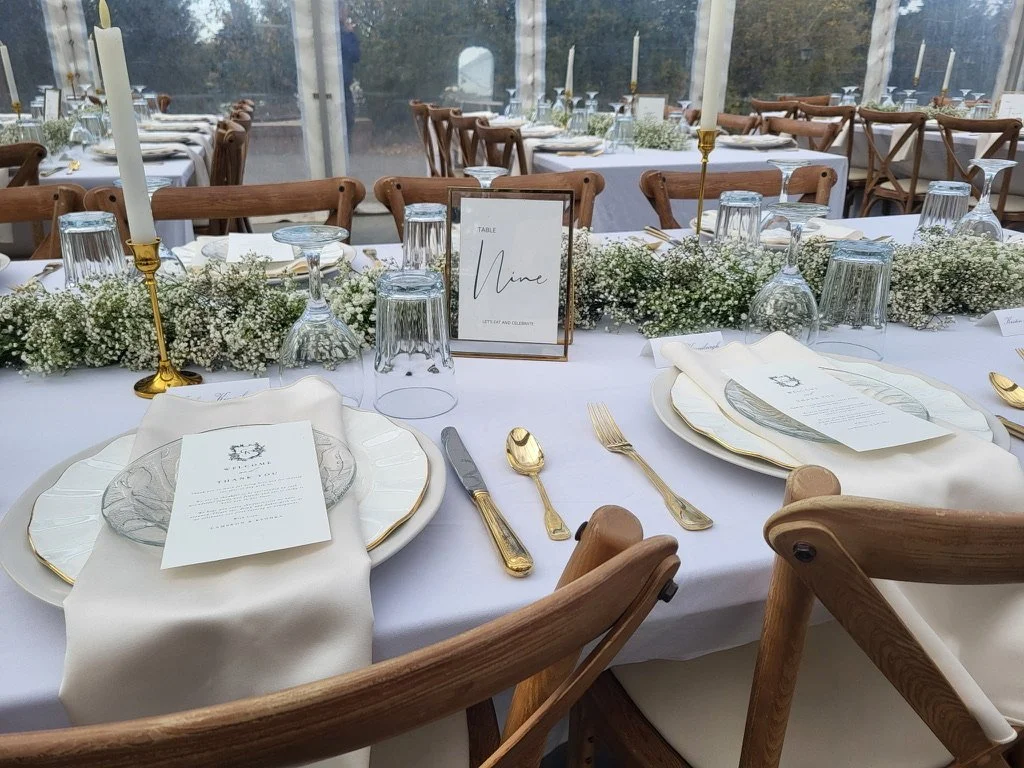 Close-up of an elegant wedding table setting inside a clear tent at Pine and Pond Wedding Venue, featuring gold cutlery, linens, candles, and greenery.