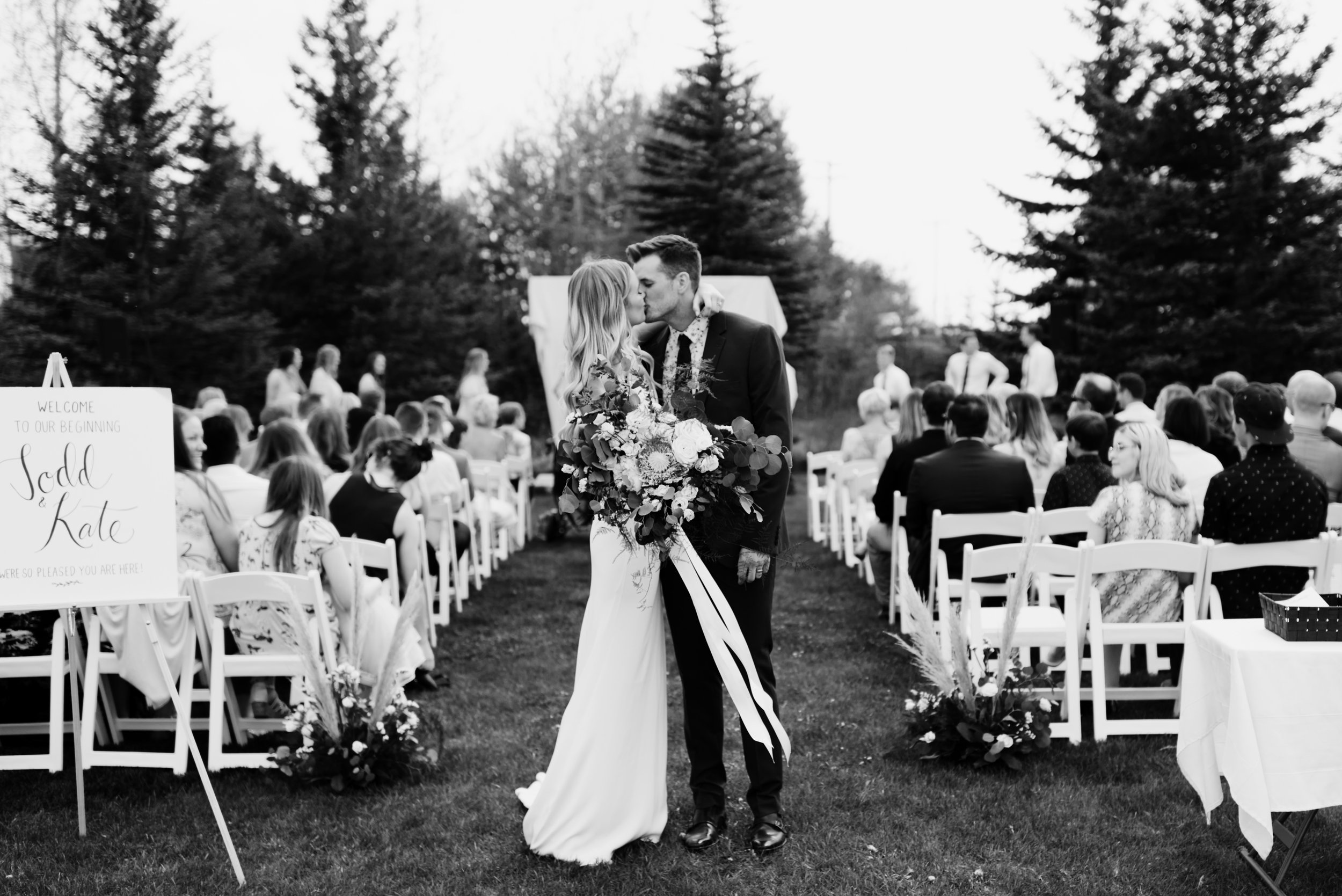 A bride and groom share a romantic kiss at the end of their outdoor wedding aisle, framed by rows of seated guests and tall evergreens. The bride holds a lush bouquet with cascading ribbons, while a personalized welcome sign and elegant décor add cha