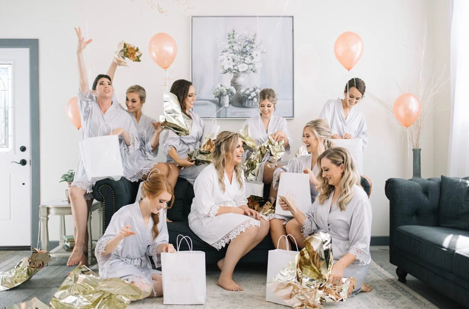 A joyful bridal party celebrates inside a bright, charming bridal dressing room located in a restored old schoolhouse. The bride and bridesmaids, all wearing matching light robes, open gift bags surrounded by peach balloons, gold wrapping, and soft n