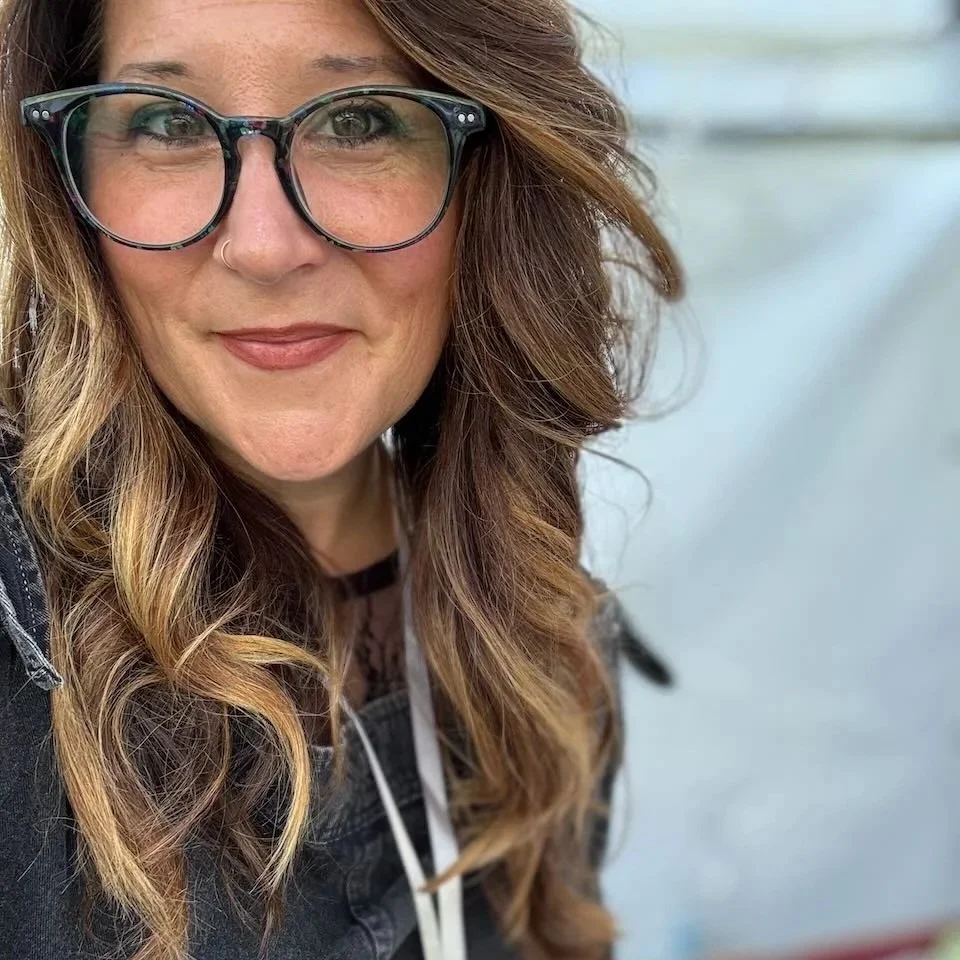 Close-up of a woman with long wavy brown hair, glasses, and a nose piercing, smiling outdoors near water.
