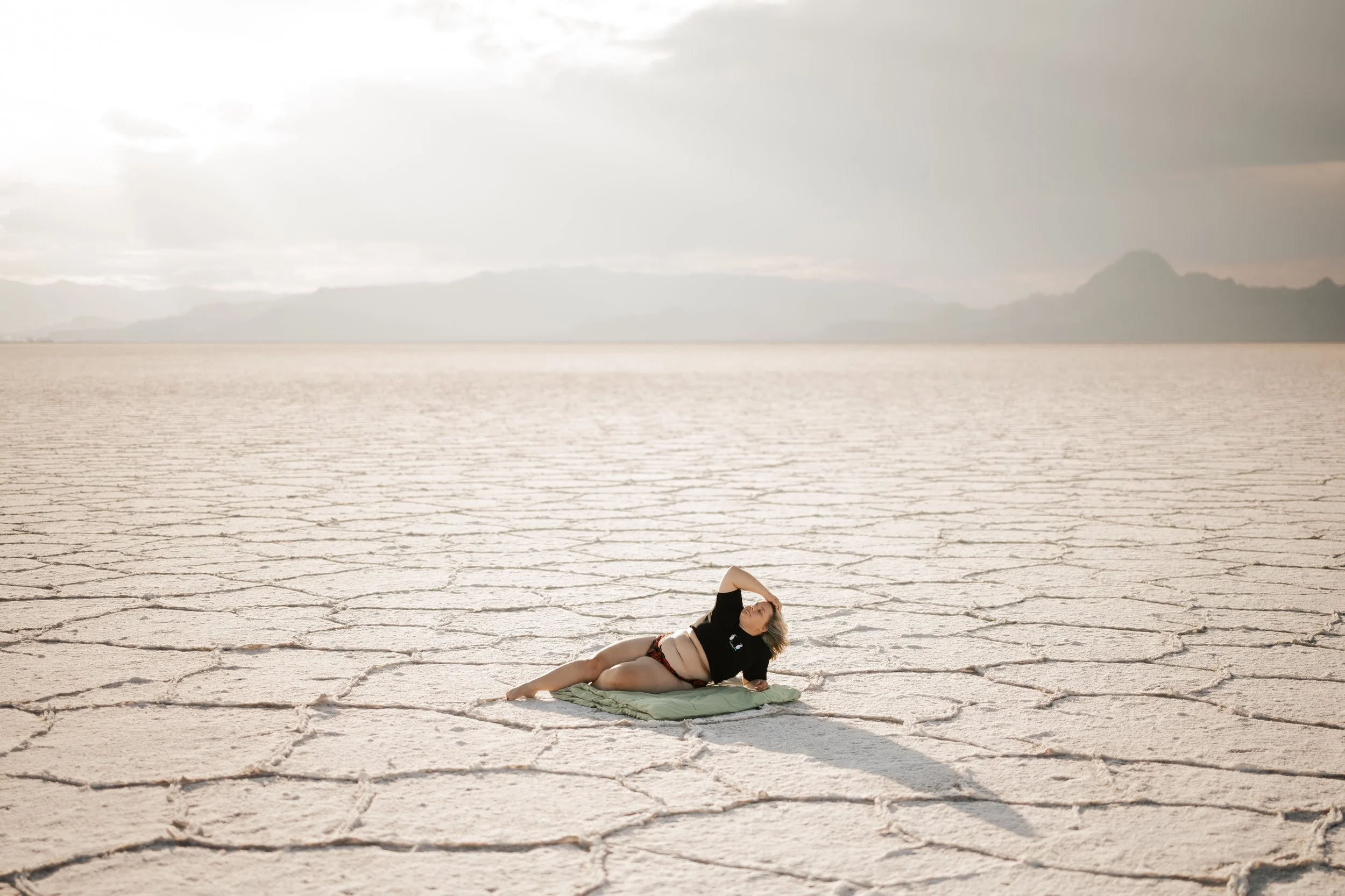 A woman lying on a blanket on cracked desert terrain under a cloudy sky.