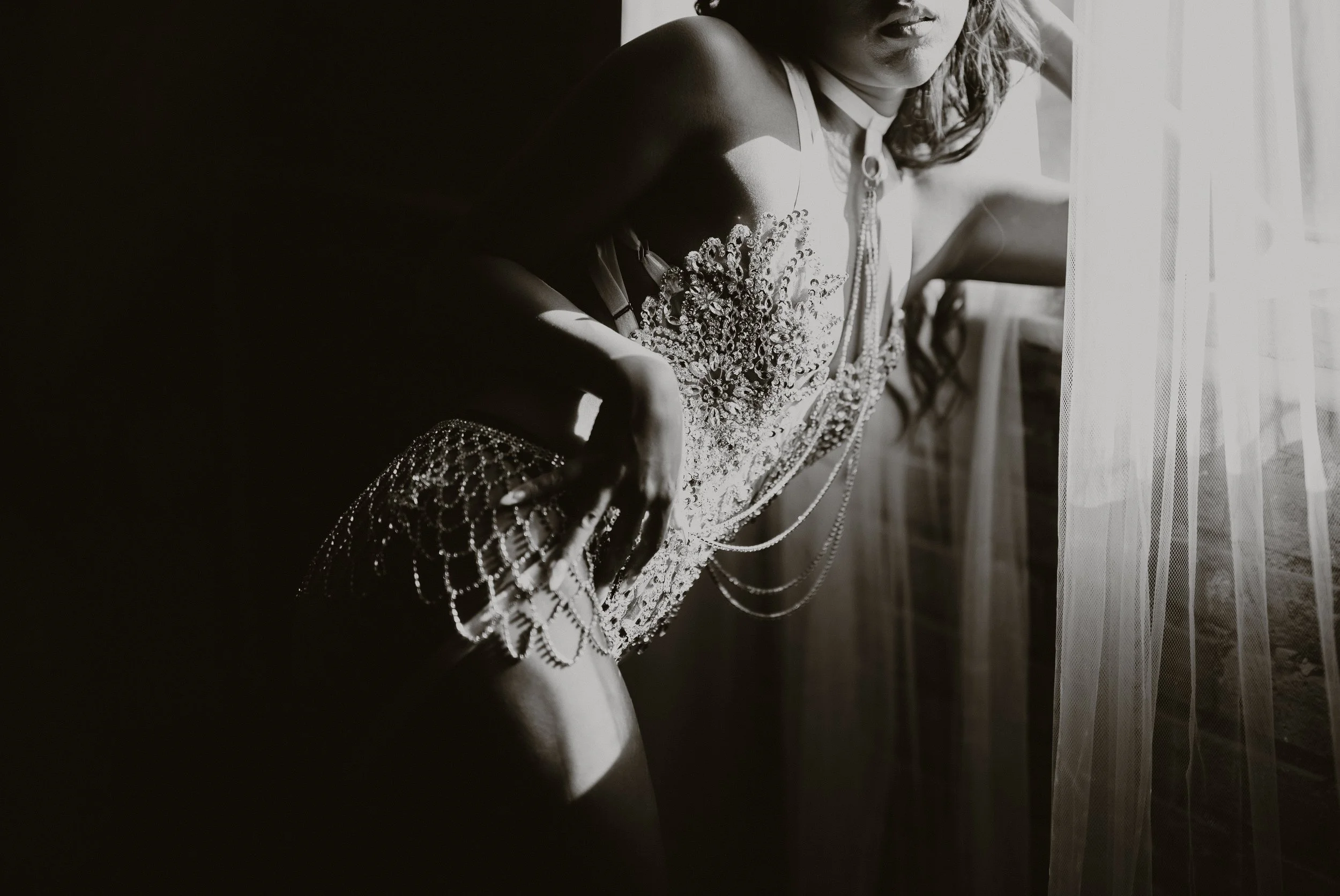 A woman in an ornate dress with intricate beadwork and jewelry, leaning near a window with sheer curtains, captured in black and white.