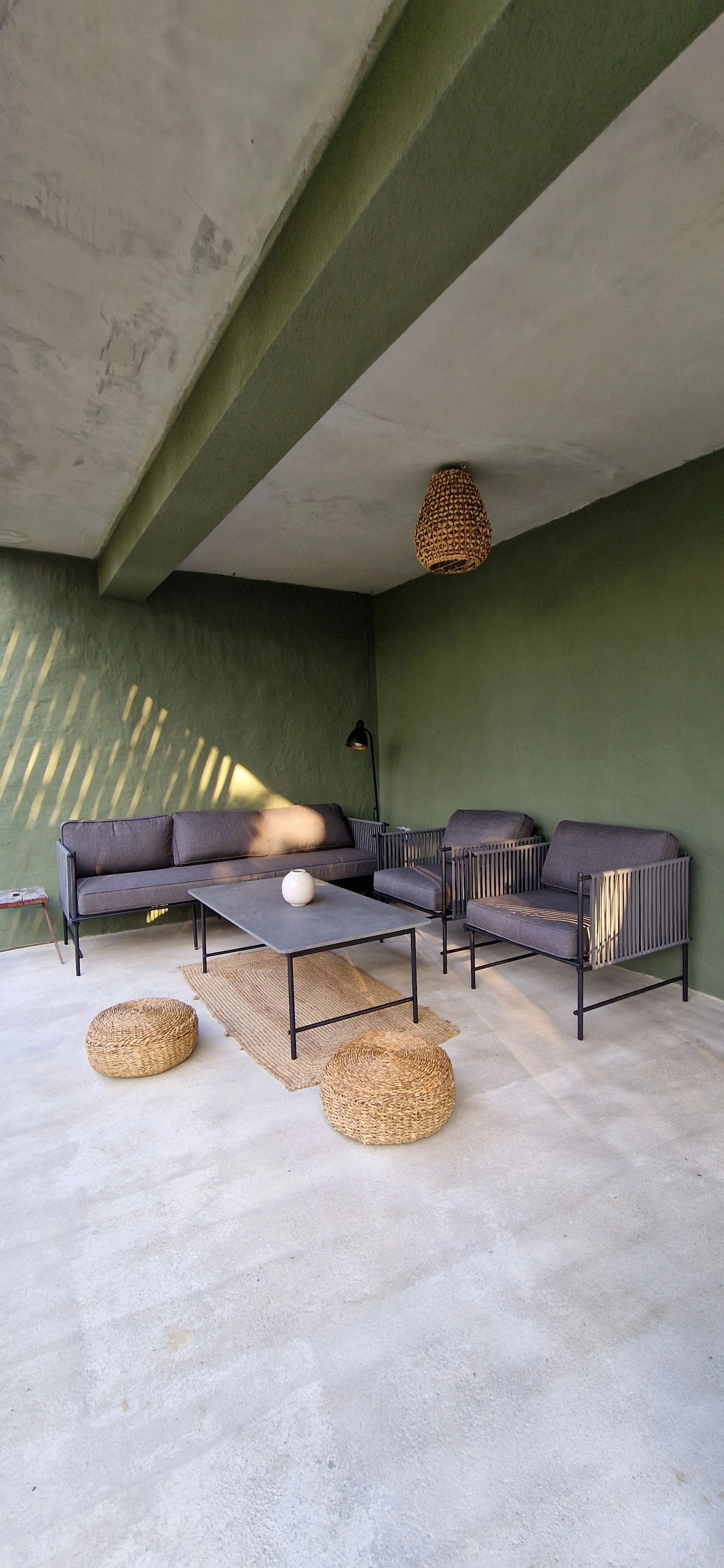 Modern living room with green walls, gray sofa, two gray armchairs, black coffee table, woven poufs, a rug, and a pendant lamp, with sunlight casting shadows on the wall.