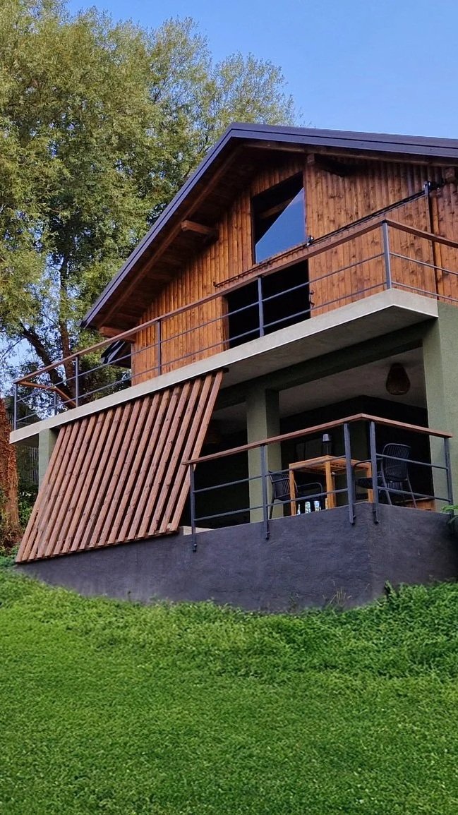 A two-story house built on a slope with an outside patio area on each level and a wooden slatted detailing on the lower level, with trees and a clear blue sky in the background.