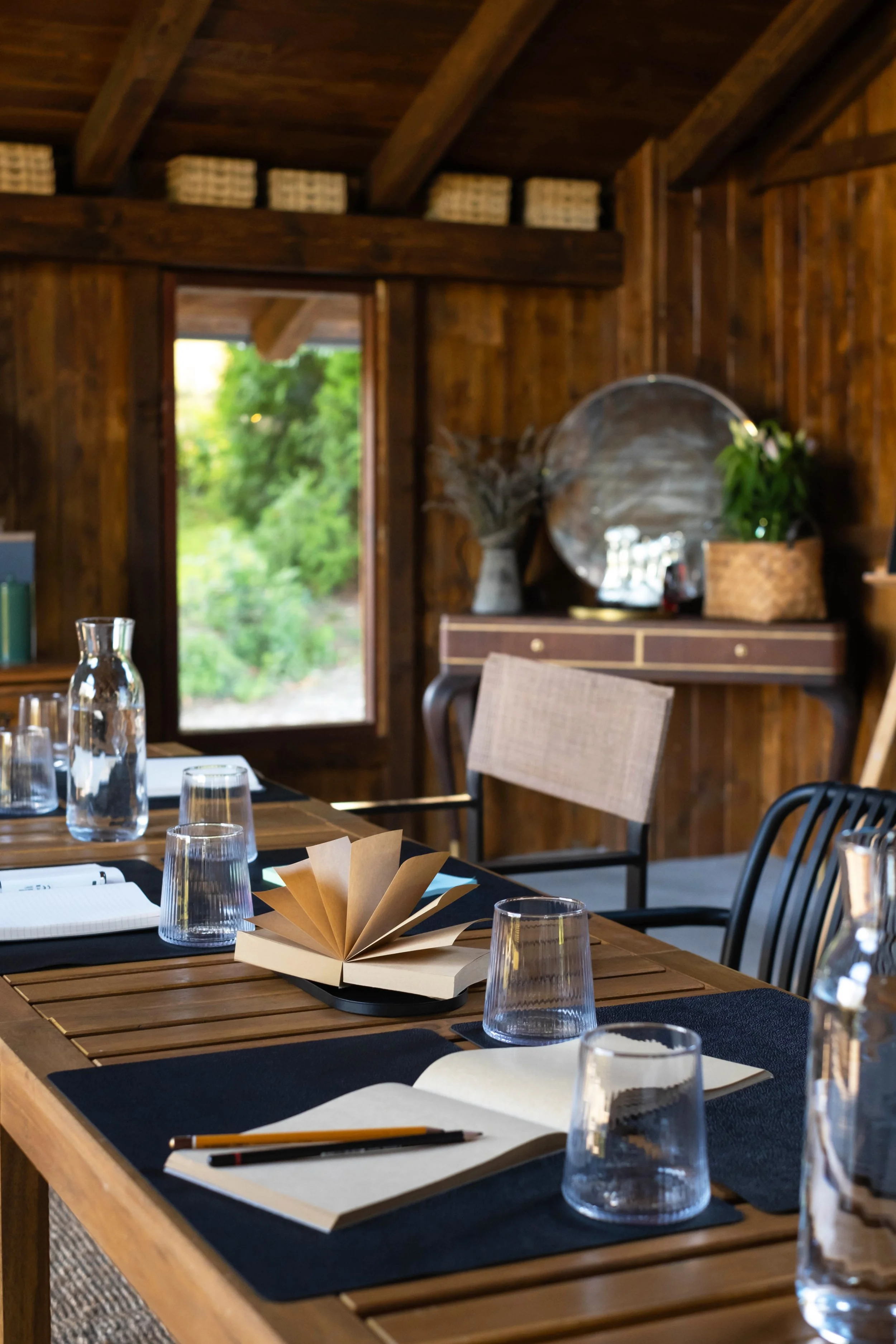 A rustic wooden dining room with a table set for a meeting or meal, featuring notebooks, pens, glasses, a water pitcher, and a decorative paper fan in the center. The room has wooden walls and ceiling, with a window showing greenery outside, and a sideboard with plants and decorative items.
