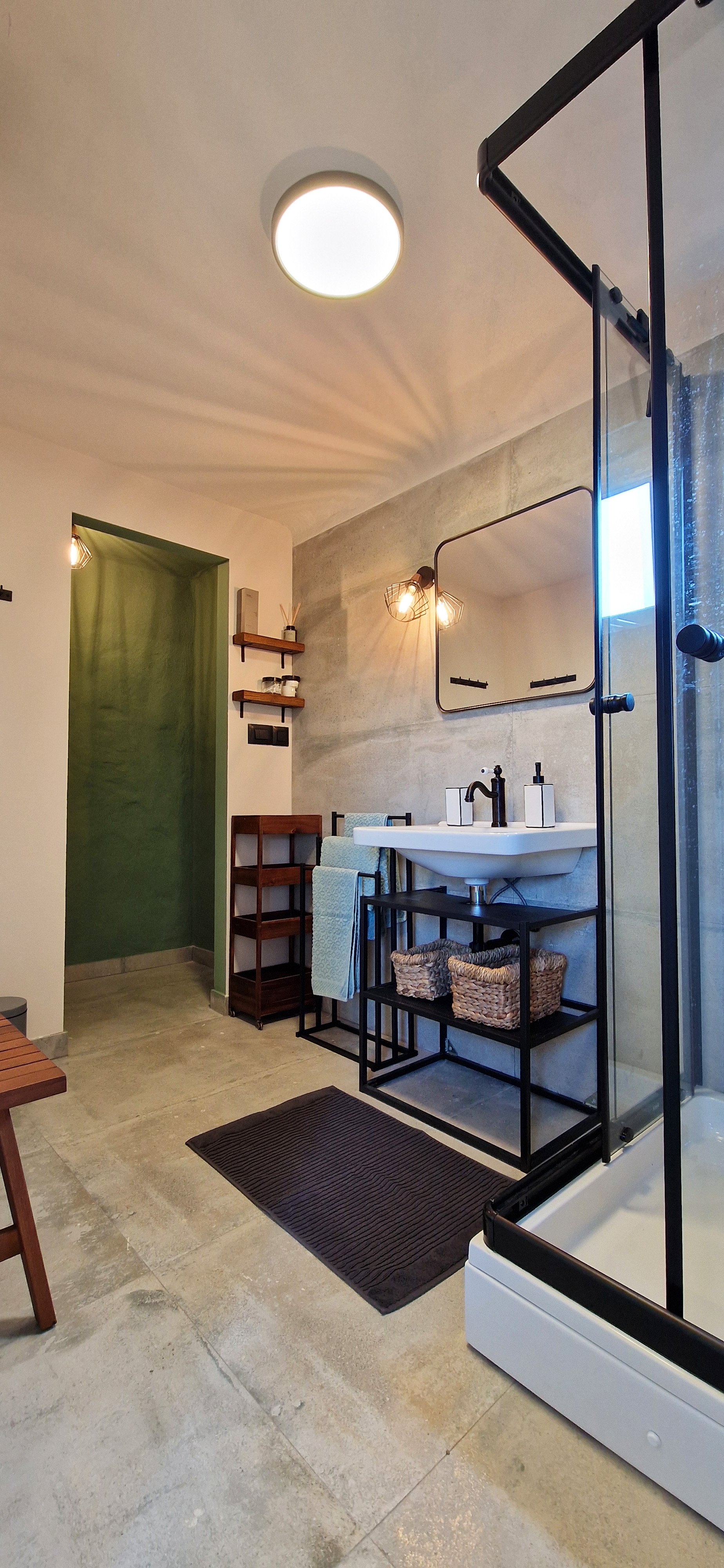 Modern bathroom with concrete walls, black metal fixtures, a white sink, wicker baskets, a black shower enclosure, and warm lighting.