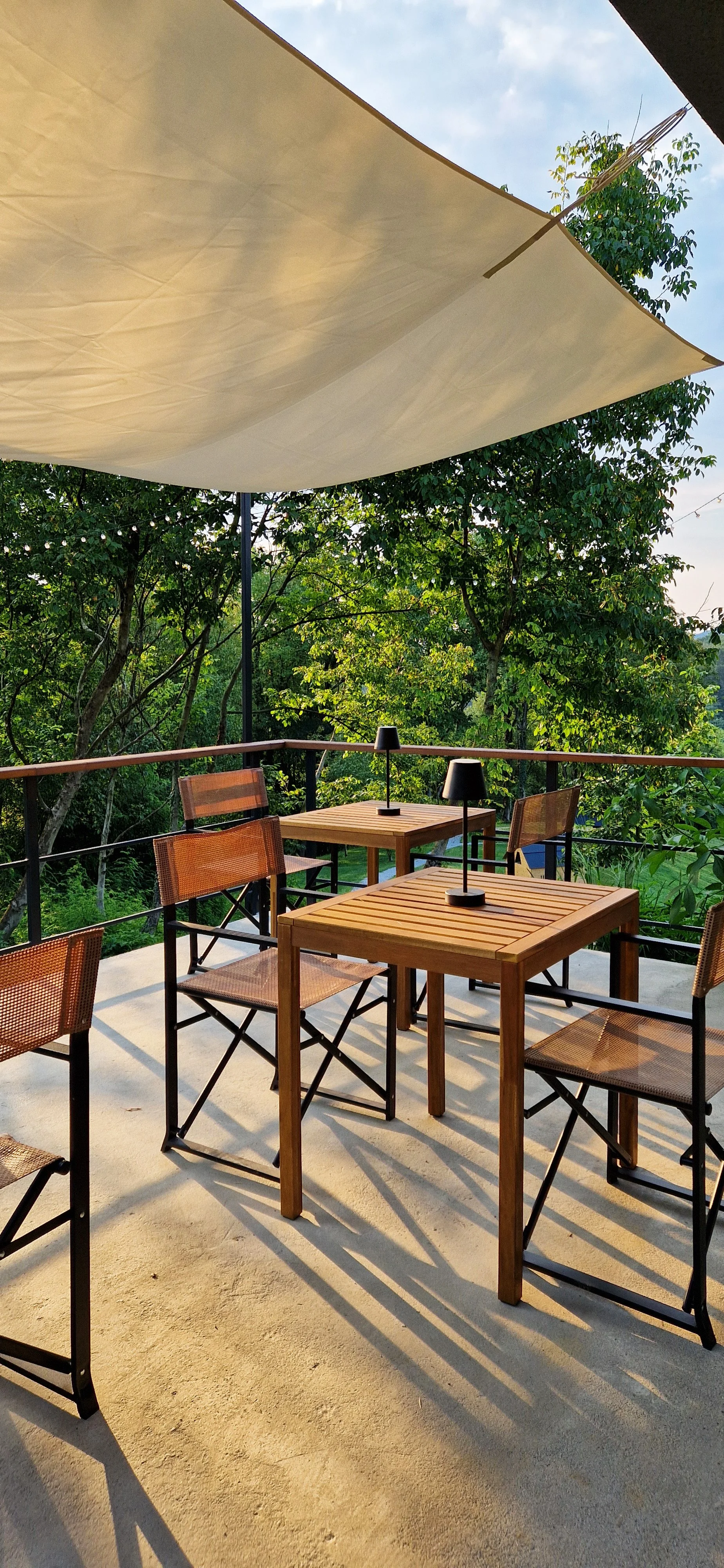 Outdoor patio dining area with four wooden tables and chairs, two small black table lamps, a large beige sail shade overhead, surrounded by green trees.