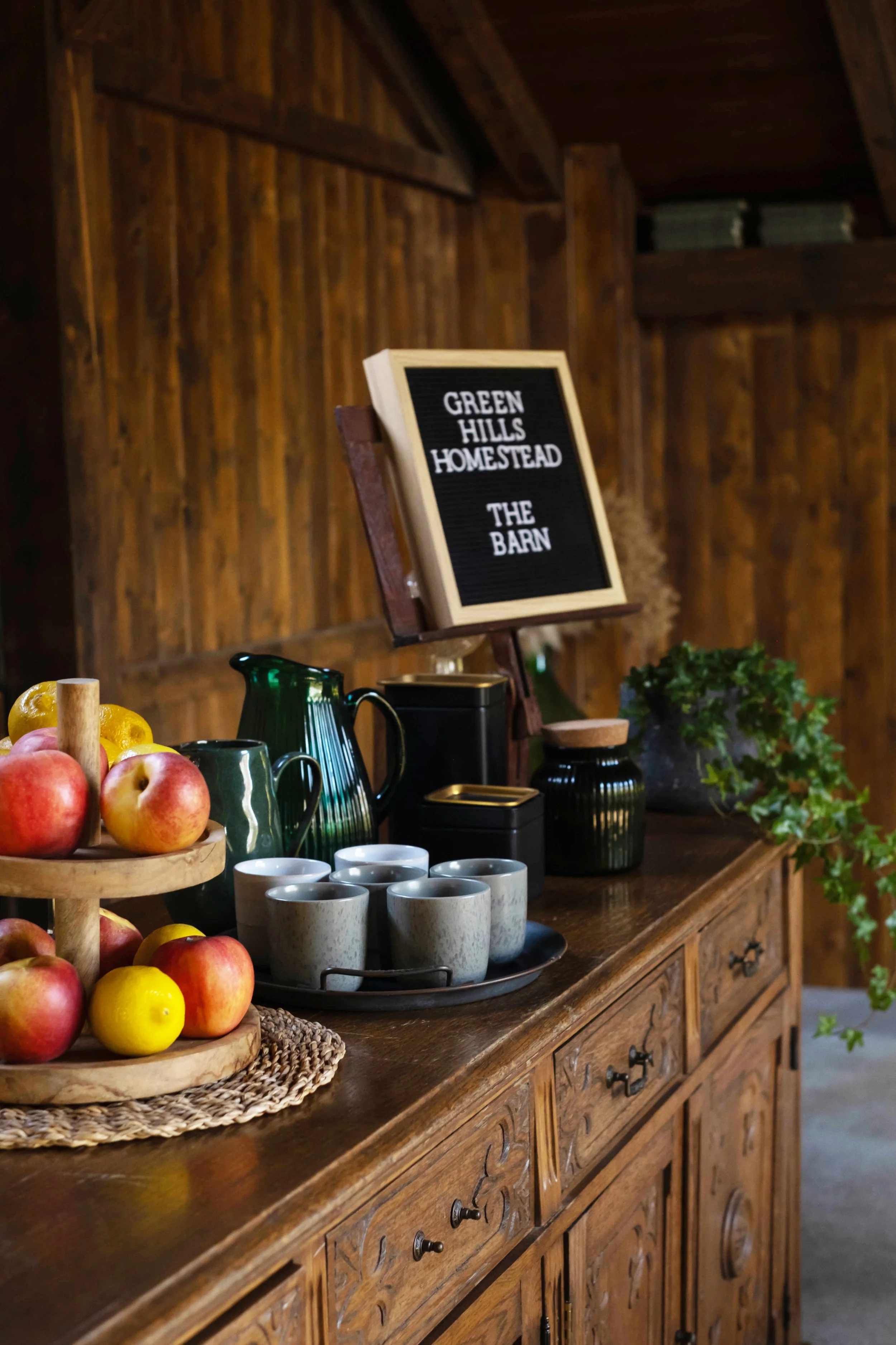 A wooden sideboard with apples, cups, and a sign that says 'Green Hills Homestead, The Barn' in a rustic setting.
