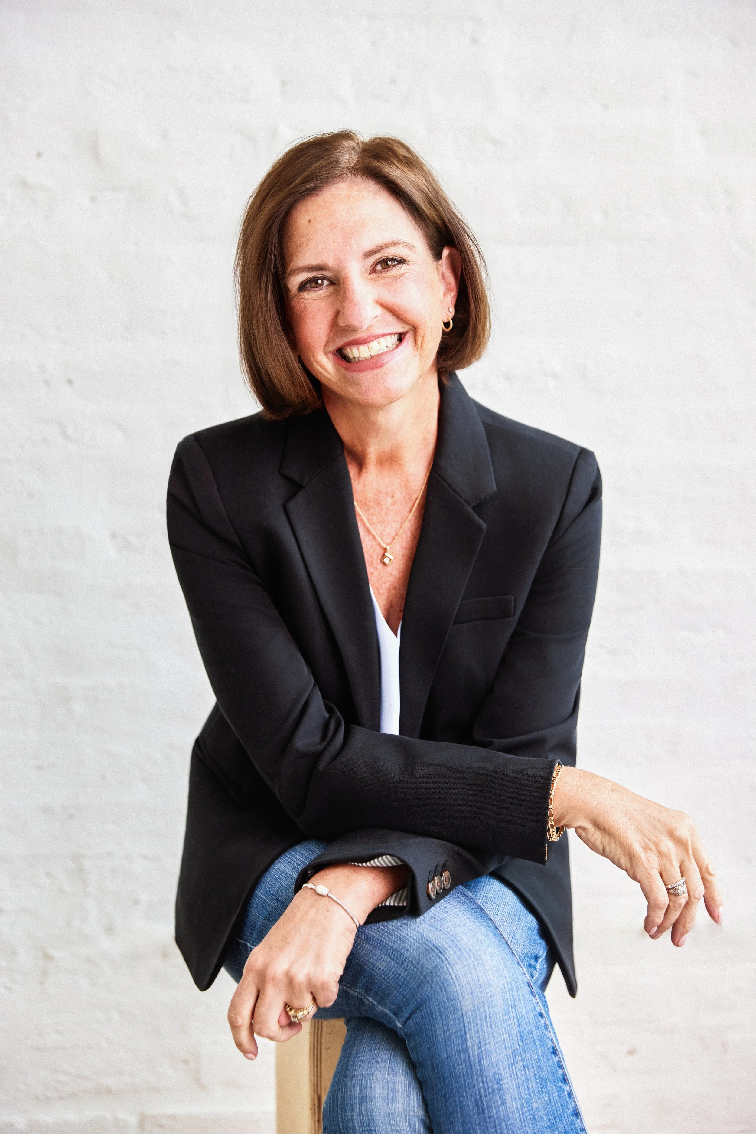 A woman with short brown hair smiling, wearing a light blue denim jacket and a light blue shirt, sitting outdoors during autumn with yellow and green trees in the background.