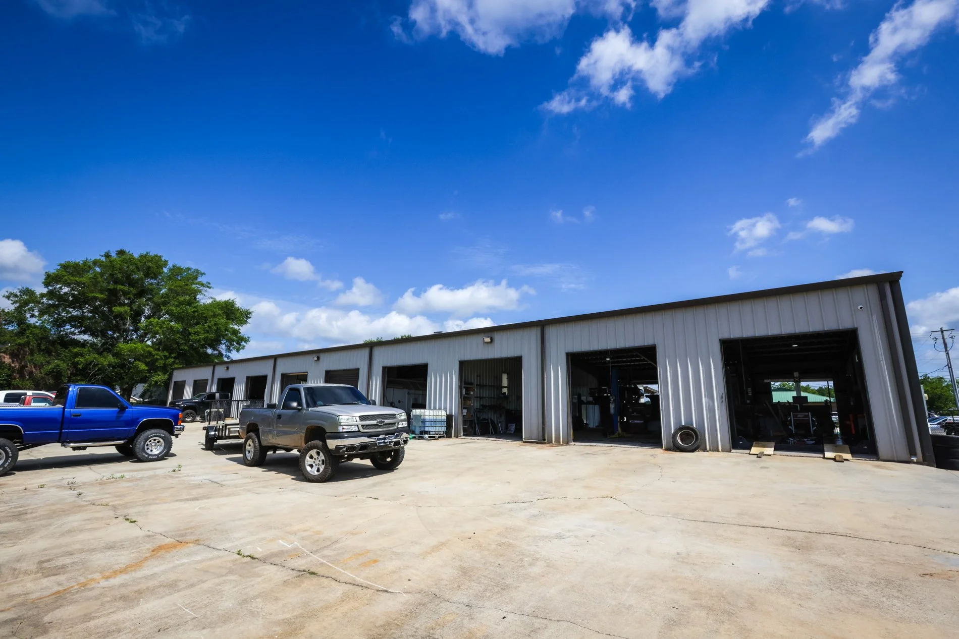 An automotive repair shop with multiple garage bays under a blue sky with scattered clouds. Several pickup trucks are parked in front of the building, and trees are visible in the background.