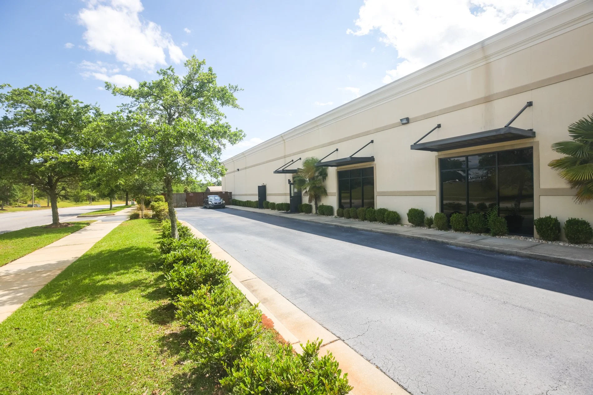 Exterior of a commercial building with large windows, black awnings, surrounded by small bushes, with a parking lot and trees in the background under a blue sky with clouds.