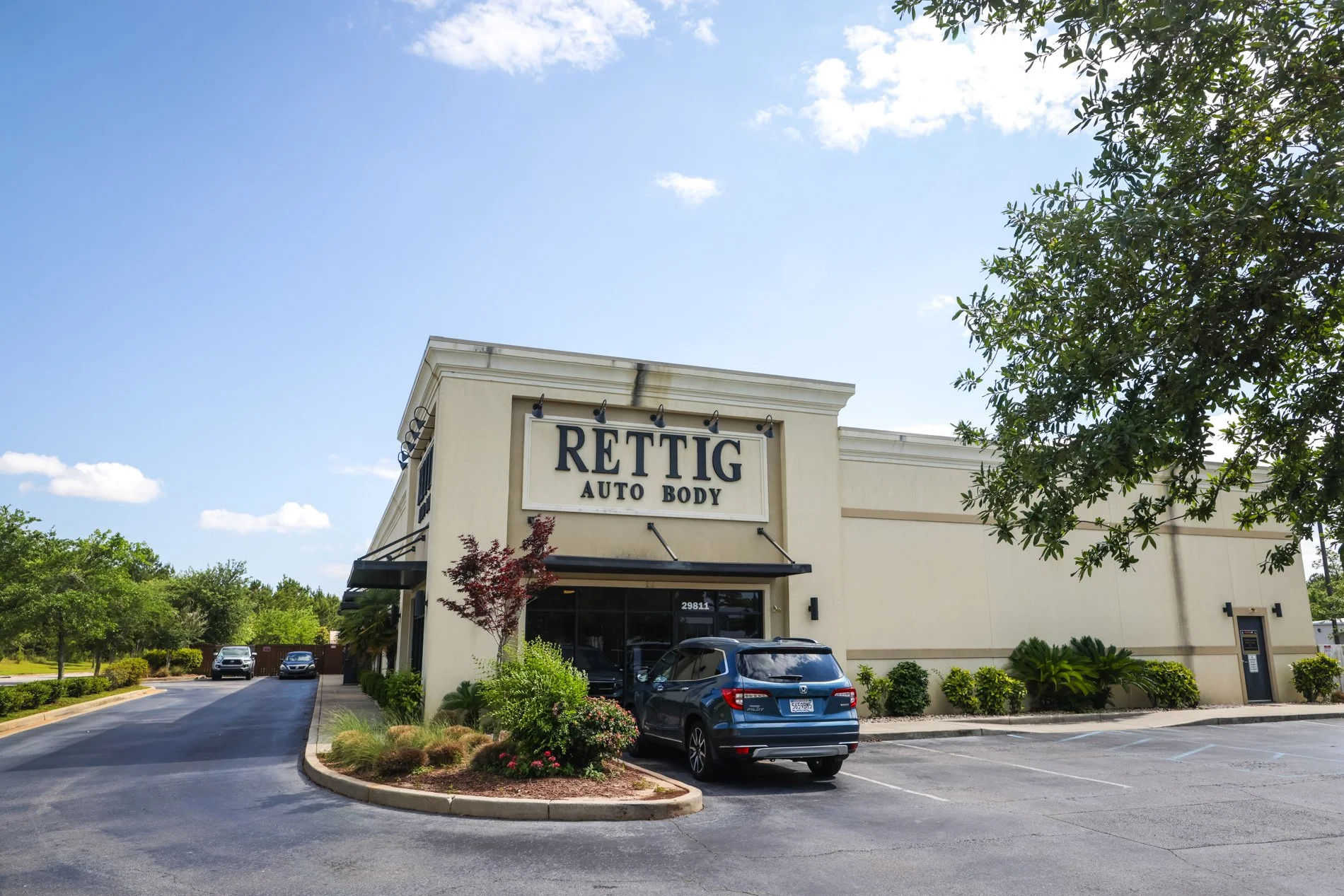 Exterior view of a beige building with the sign 'RETTIG AUTO BODY' above the entrance, parked cars in front, landscaped greenery, trees, and a clear sky with some clouds.