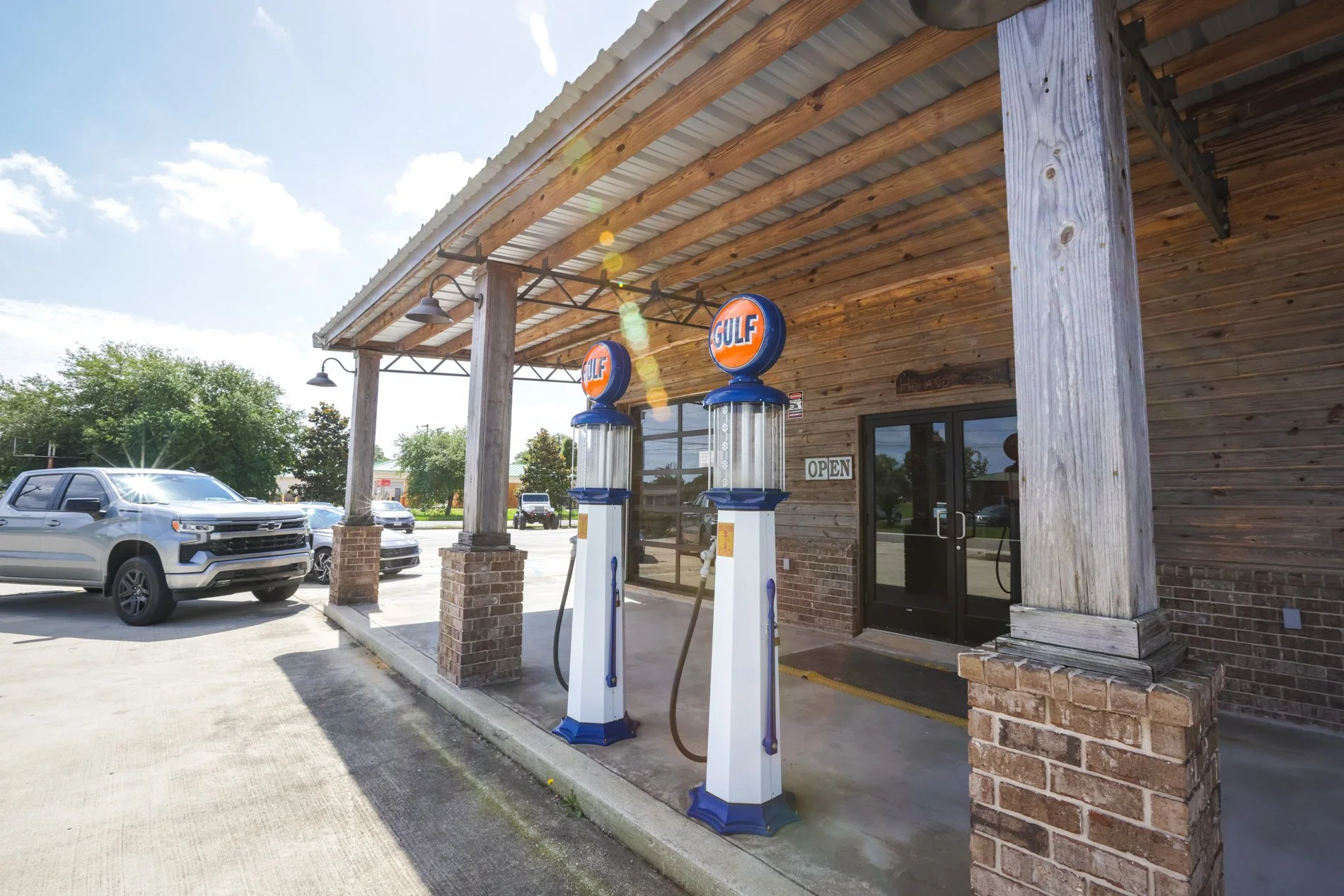 Exterior of a small gas station with two vintage Gulf gasoline pumps under a wooden roof, parked cars in the lot, and a sign indicating the station is open.