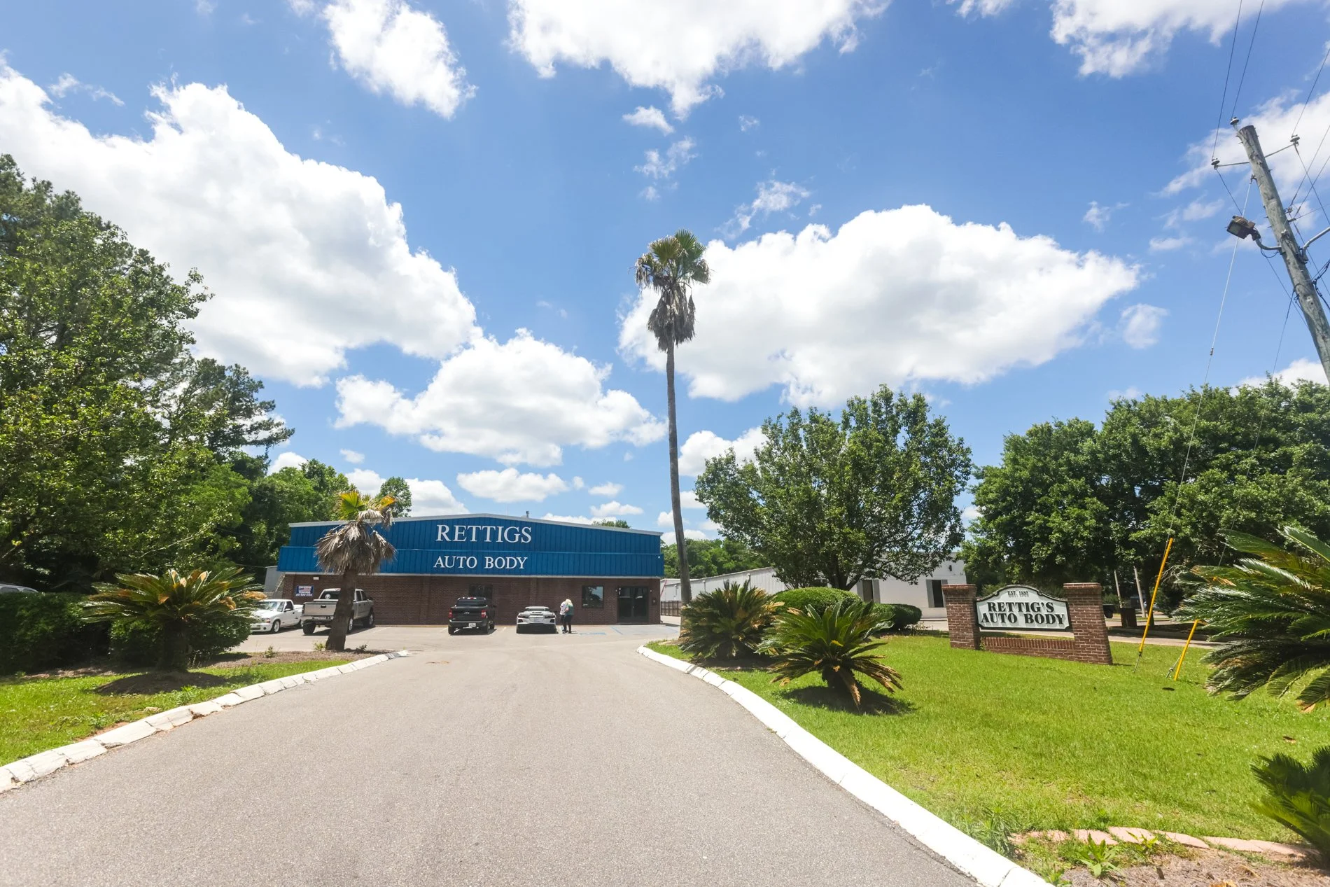A parking lot in front of Rettig's Auto Body with several cars parked, surrounded by greenery including trees and bushes. A tall palm tree stands in the center, with a blue sky and scattered clouds above. There is a sign with the auto body shop's name on a brick pillar and a blue building with signage.