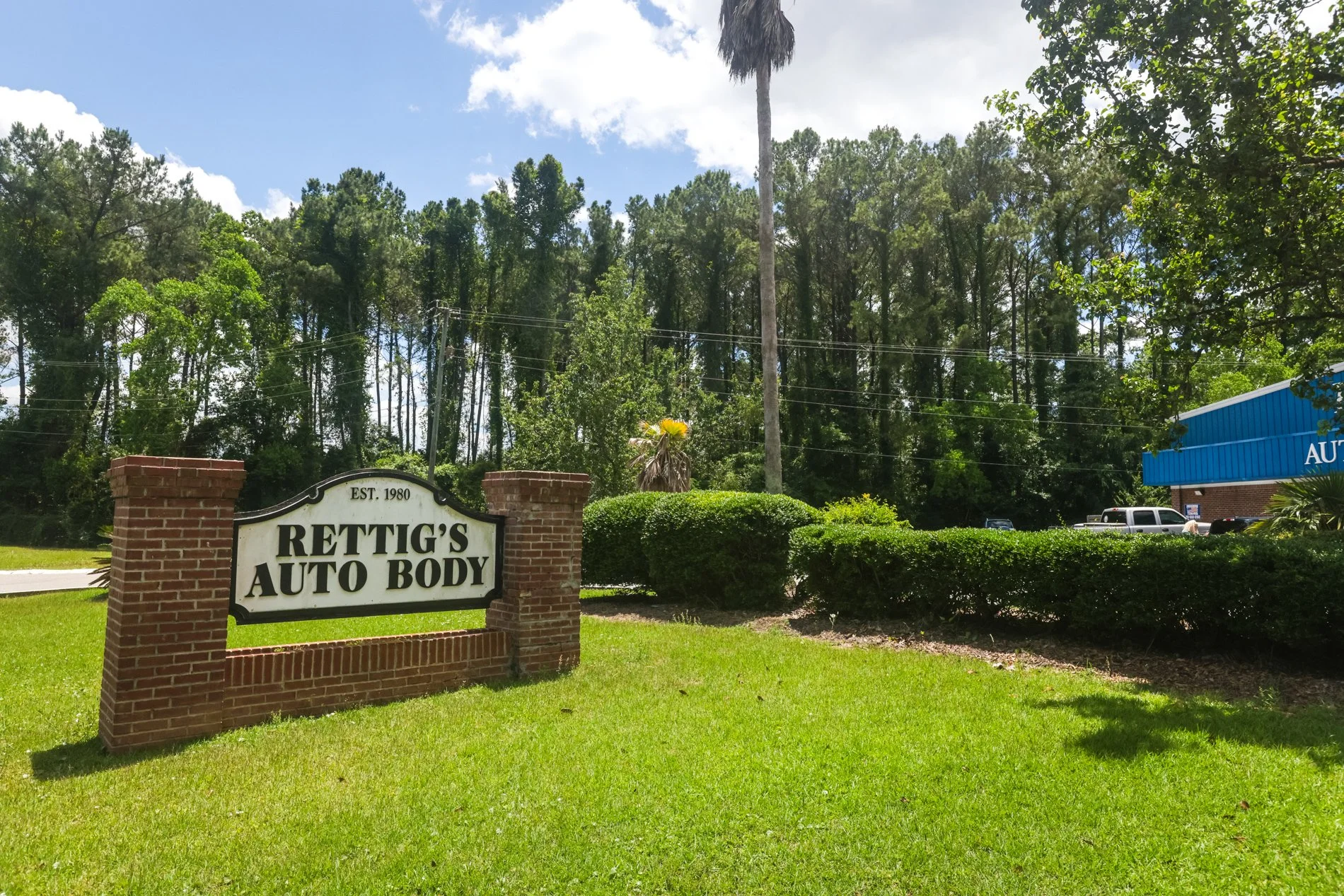 A brick sign reads 'RETTIG'S AUTO BODY EST. 1980' in front of a lush green lawn. Behind it, tall trees and a utility pole with power lines are visible under a partly cloudy sky. Part of a blue building and parking lot are on the right.