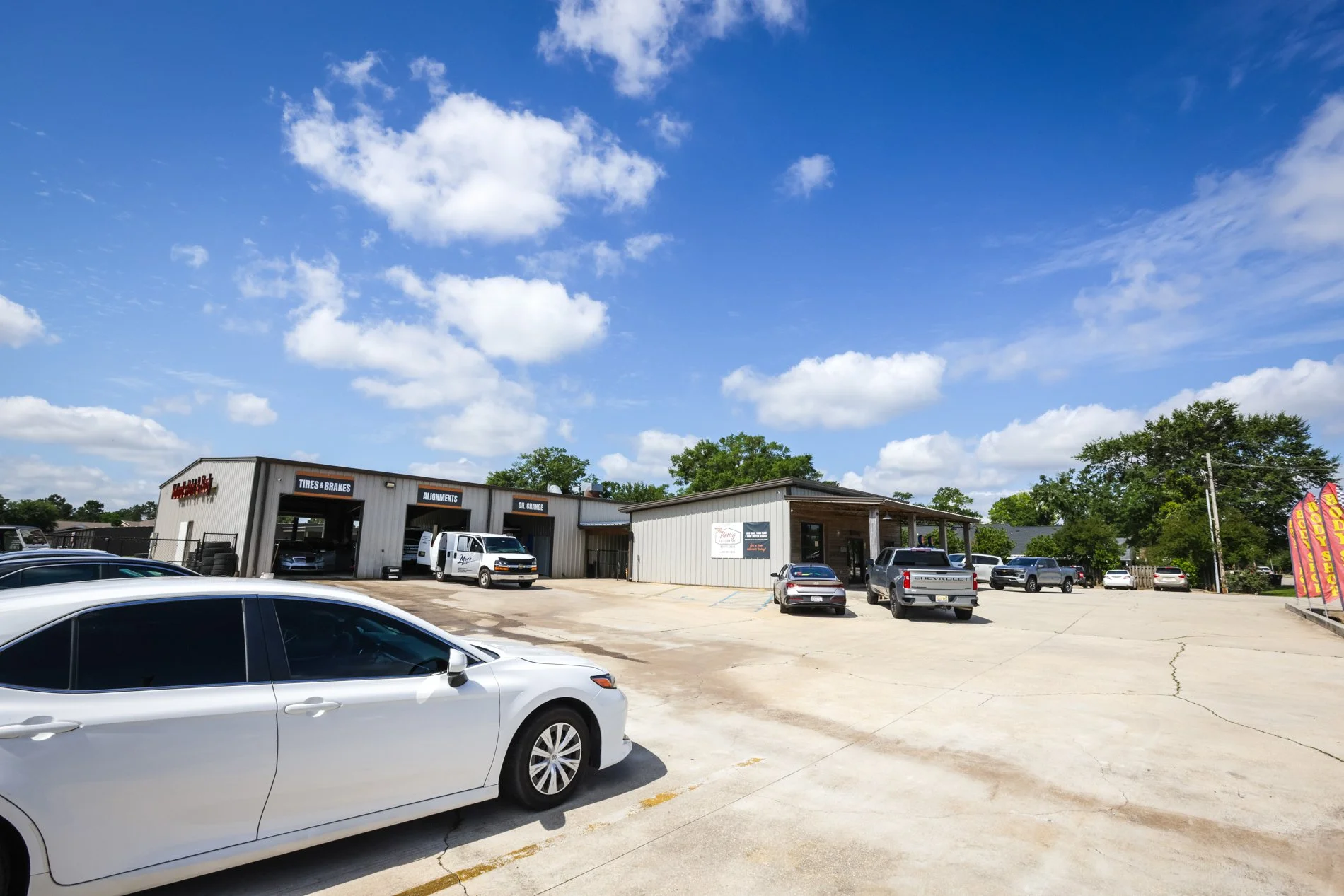 Auto repair shop with multiple service bays, parking lot with cars, clear blue sky with scattered clouds, and trees in the background.