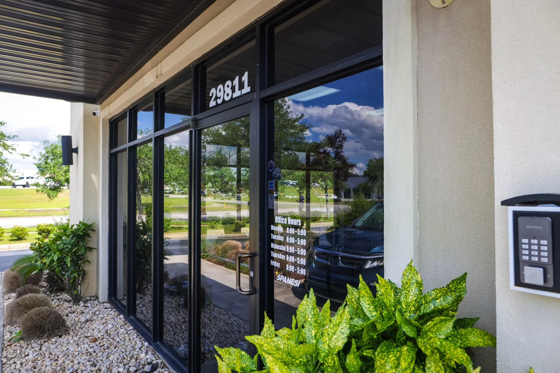 Exterior view of a building with glass storefront, displaying business hours on the window. The building has a modern design with beige walls, black framing, and a dark wood soffit. There's a landscaped area with green plants and rocks, and a parking lot with cars visible in the reflection of the glass.