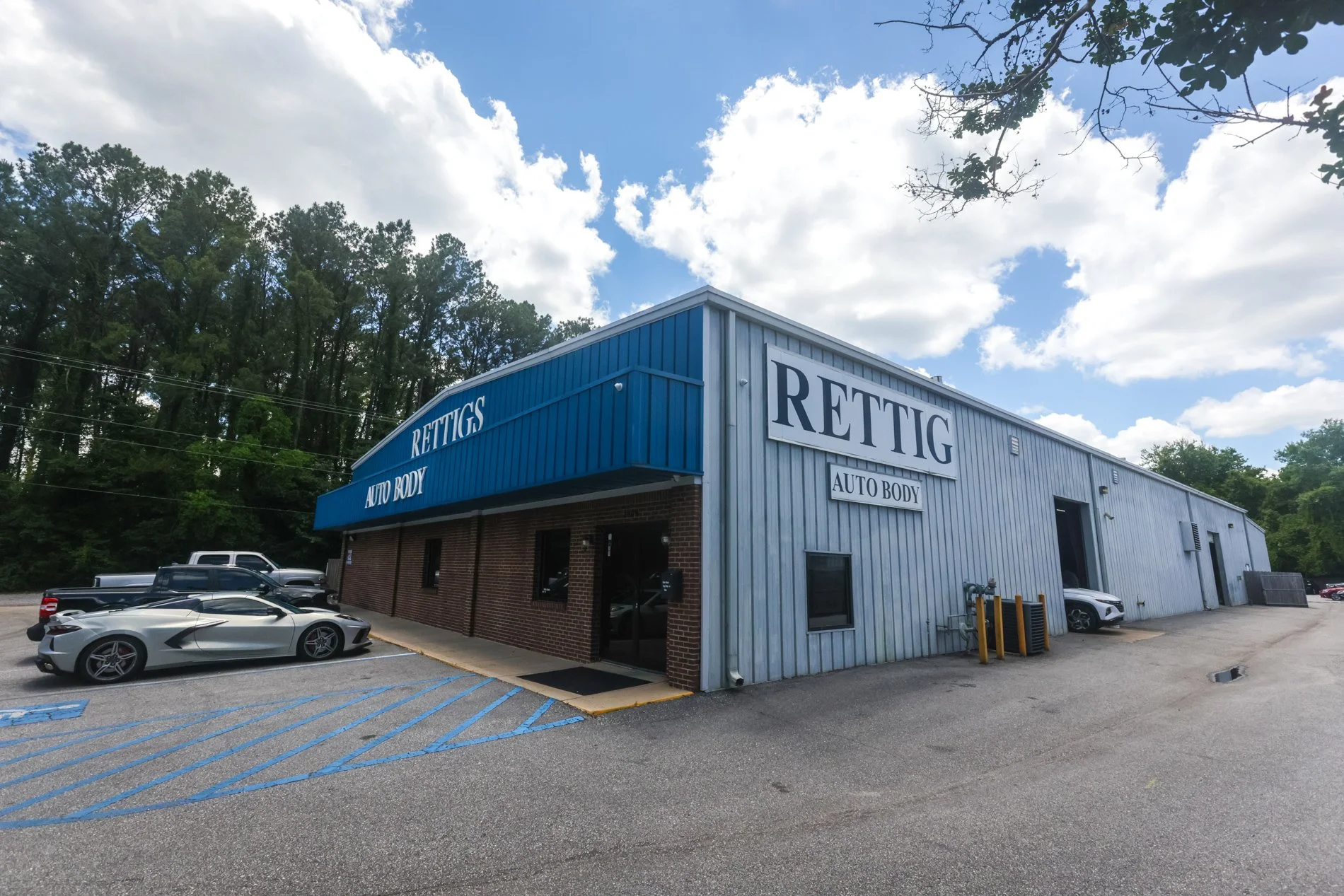 Exterior view of a blue and brick auto body shop called 'Rettig Auto Body' with parking lot, several parked cars, and a partly cloudy sky.